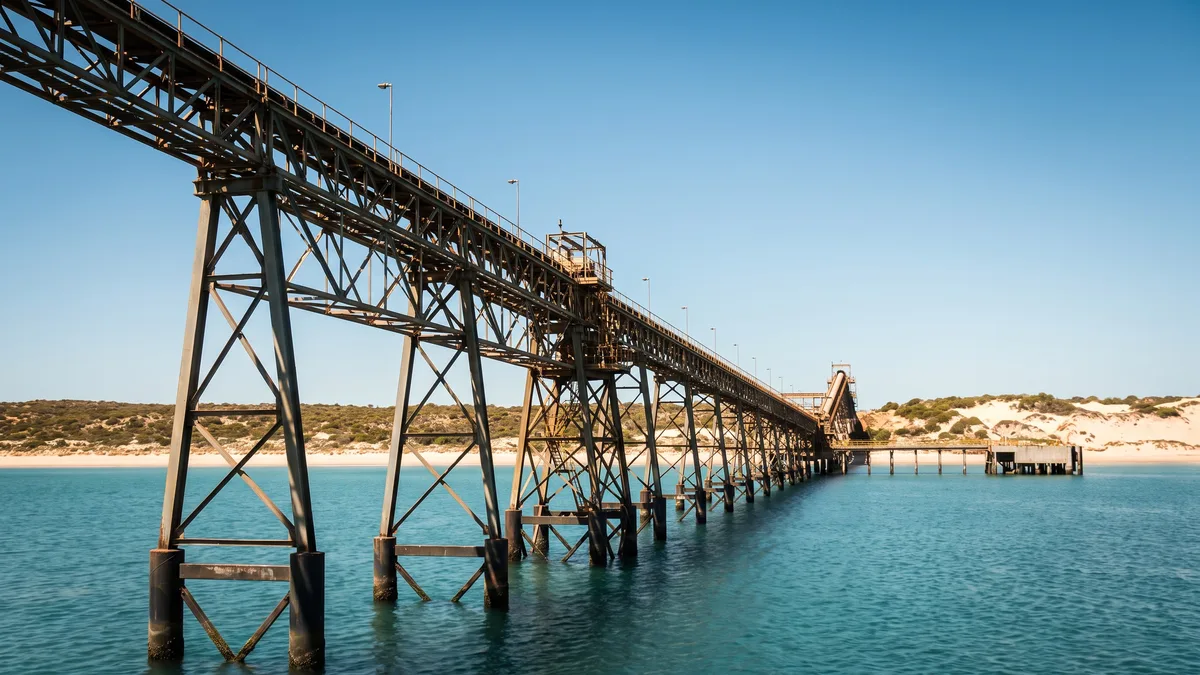 A long trestle conveyor jetty over calm turquoise water, with low arid coastal scrub and pale dunes behind.