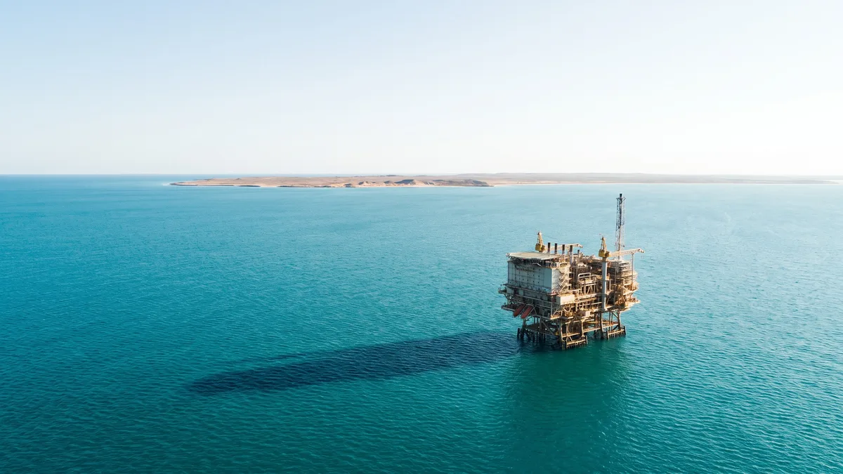 A distant offshore gas processing platform on open turquoise ocean, with a low arid island coastline on the horizon.