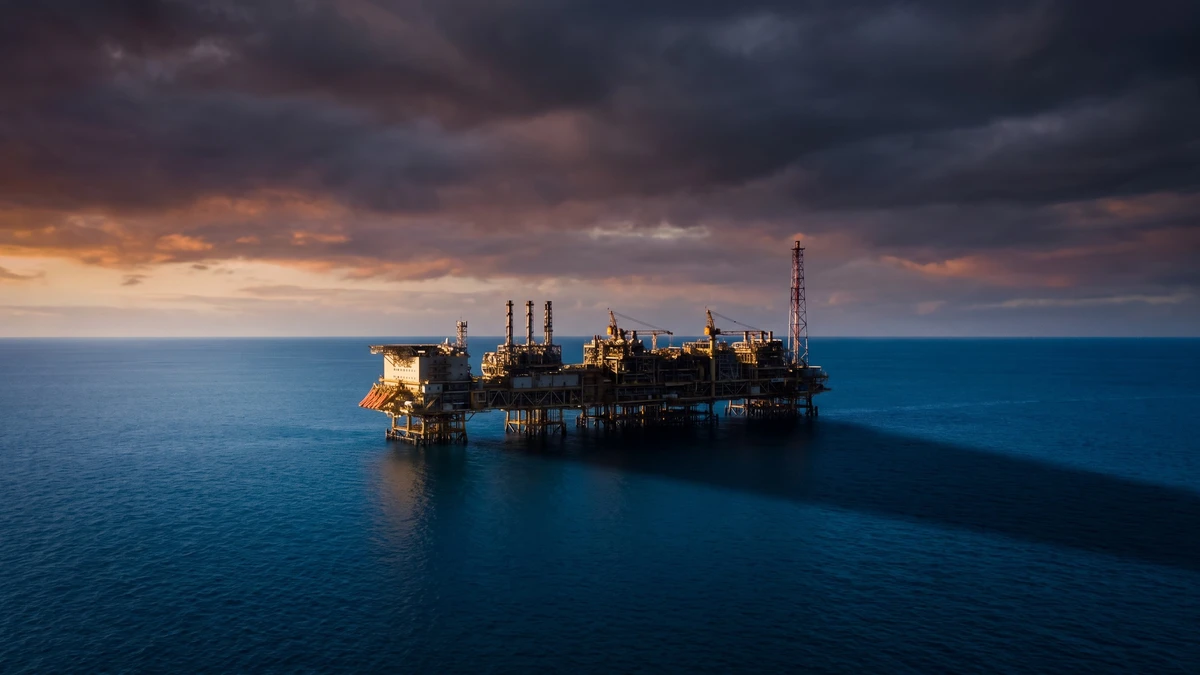 A distant offshore gas processing platform on open ocean at dusk, under a dramatic sky.