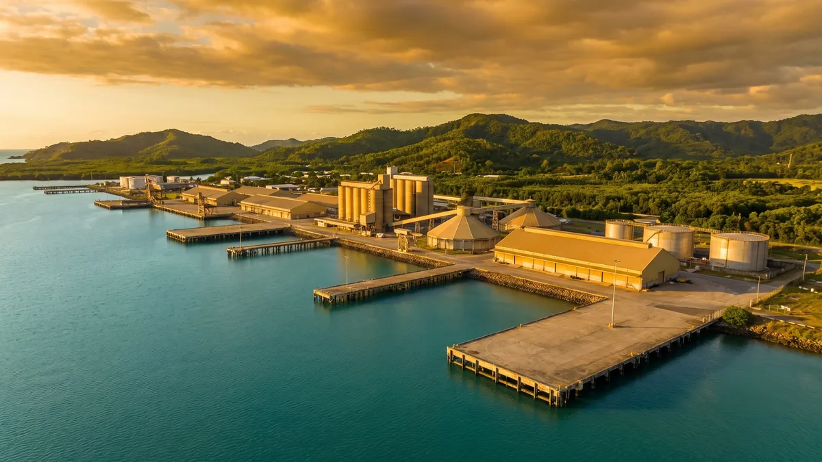 An elevated dusk view of a multi-wharf harbour with grain silos, sugar and bulk storage sheds and fuel storage tanks, calm turquoise water and tropical hills behind.
