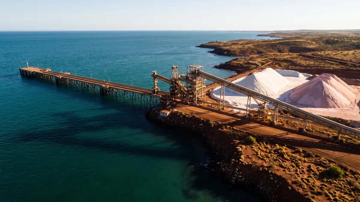 A bulk-minerals conveyor loading jetty on the open Pilbara coast, with pale white and pink salt and potash stockpiles beside the loader, dark turquoise tidal water, and arid red-earth coastal terrain with sparse spinifex scrub behind.