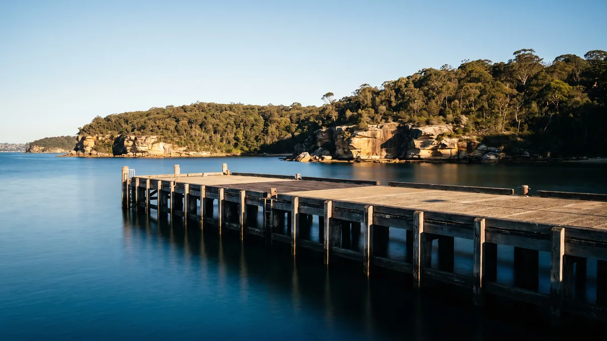 A passenger and cruise terminal wharf on calm Sydney Harbour water, with sandstone headlands and temperate bushland behind, no cargo or containers.