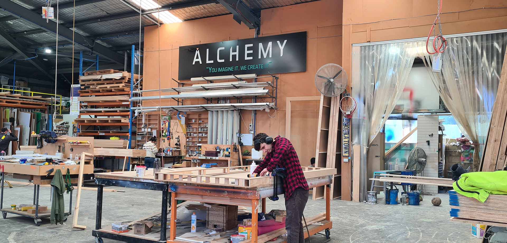 Alchemy workshop factory space with tools and timber on shelves and a man with a checked shirt screwing in screws with a drill
