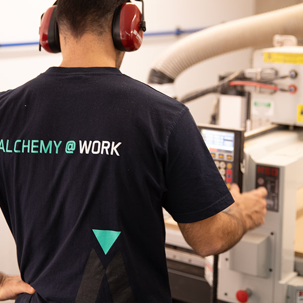 worker from behind wearing alchemy branded tee shirts operating the CNC machine