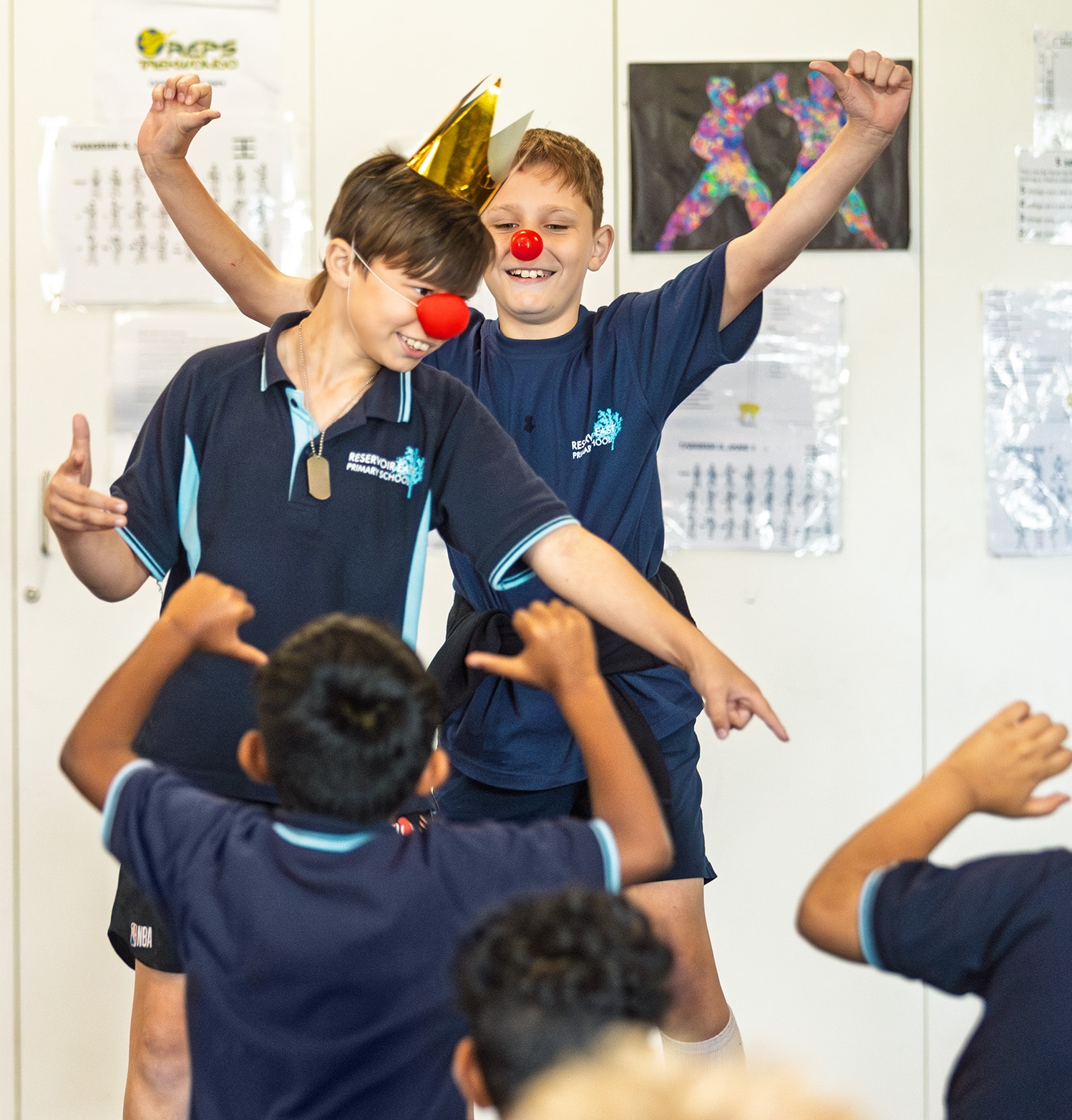 two primary aged boys are performing enthusiastically for their classmates. they wear red clown noses and one has a gold paper crown. one has his arms in the air, the other is pointing into the crowd. the audience have their arms in the air making a "thumbs down" gesture.