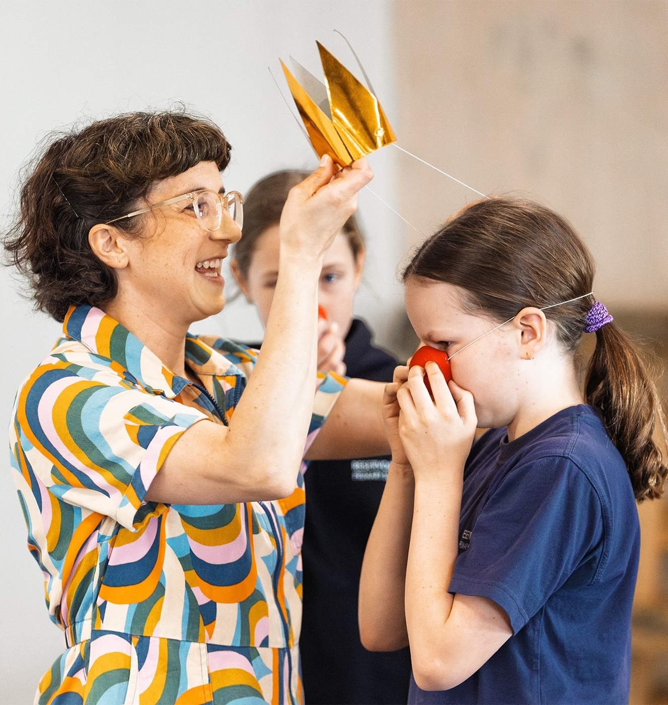 Debbie Zukerman is helping a primary aged girl put on a gold crown. the girl is putting on a red nose, getting ready to perform for classmates.