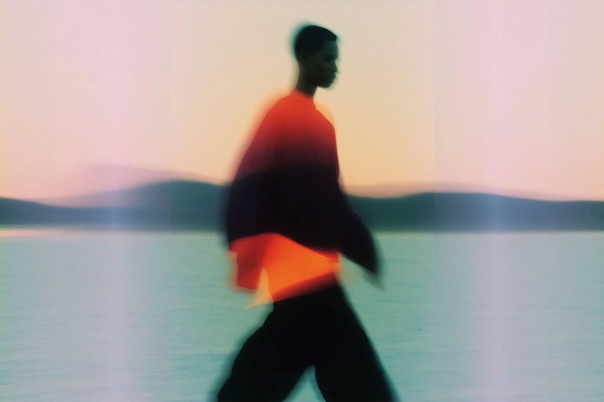 Blurred silhouette of a person walking near water during sunset with hills in the background.