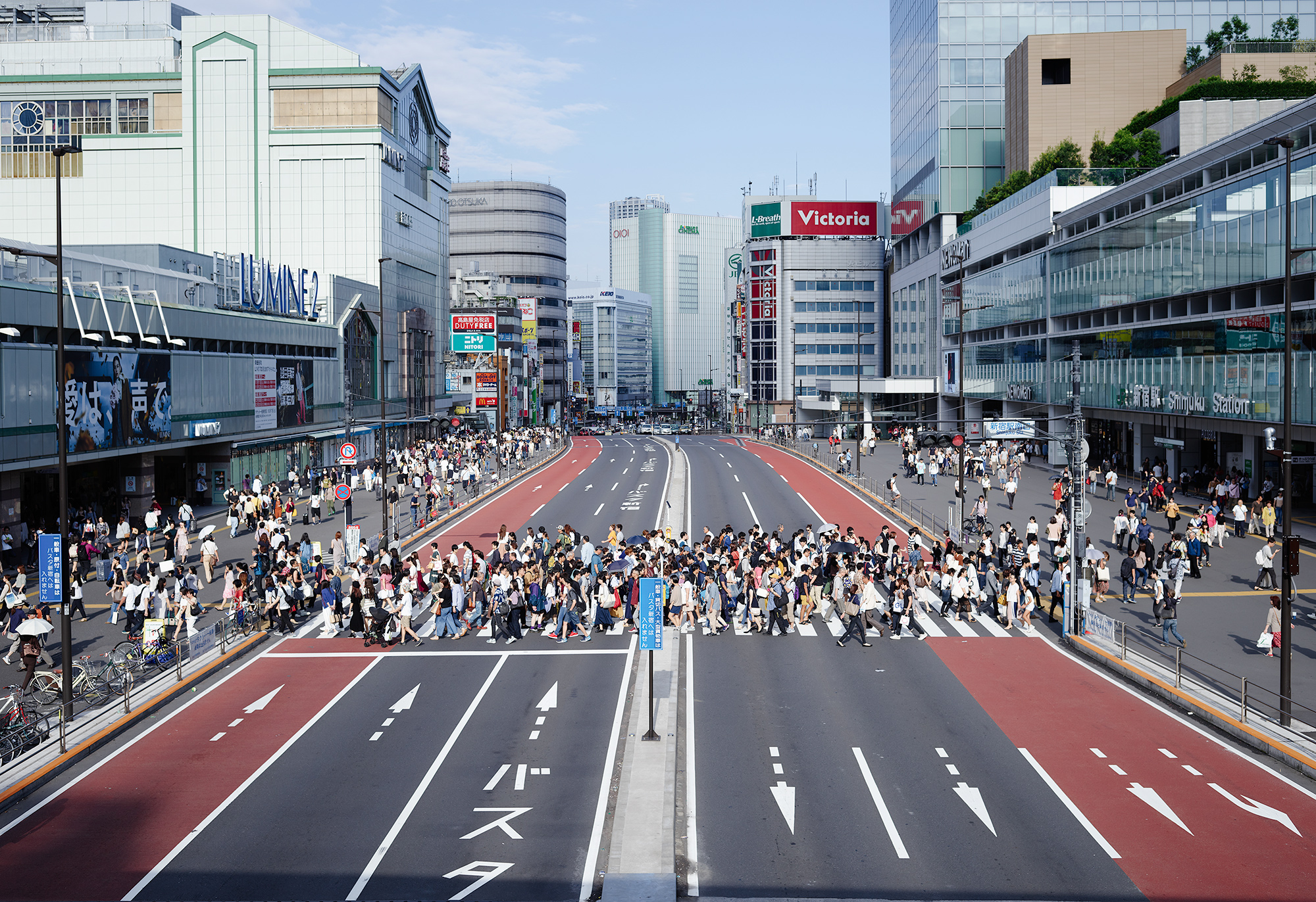 Crowded pedestrian crosswalk at Shinjuku Station in Tokyo with people crossing wide streets surrounded by tall buildings and shops.