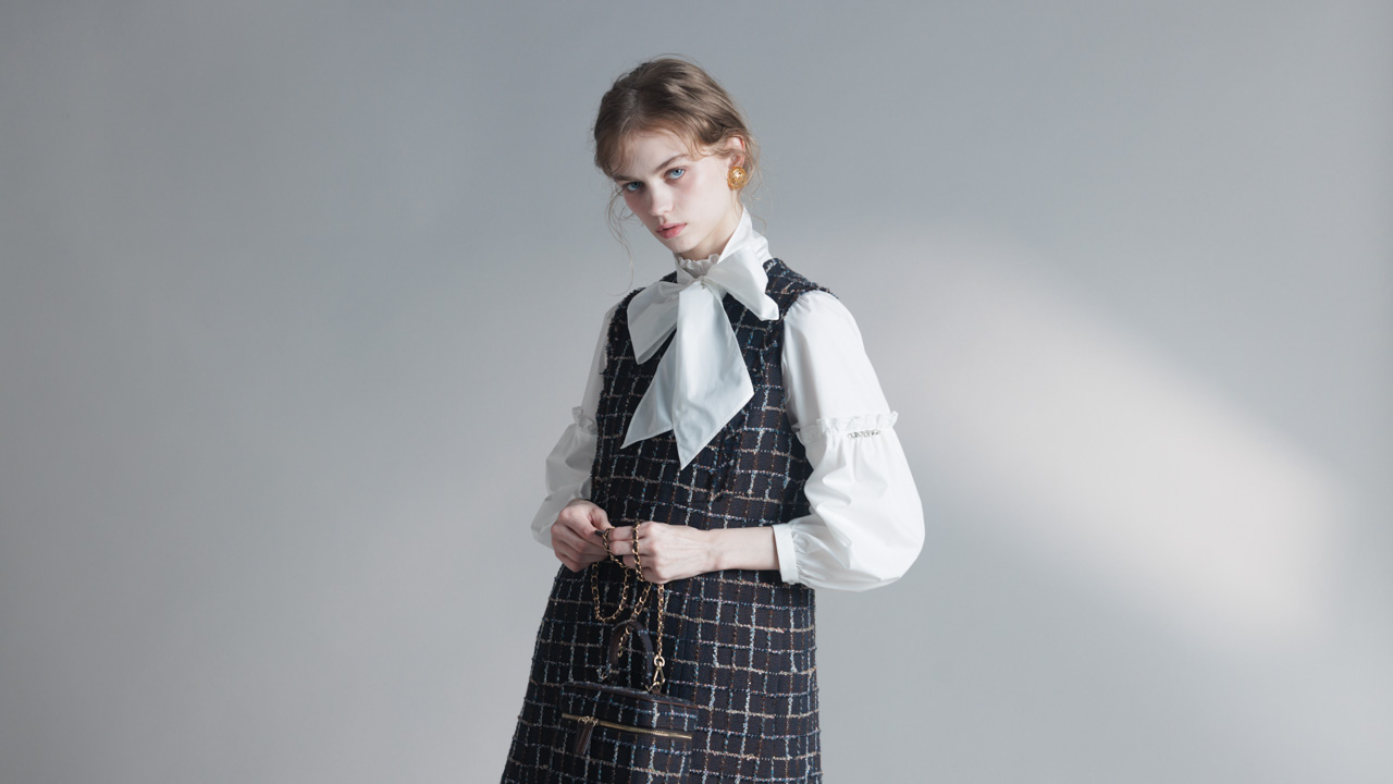 Young woman wearing a black checkered dress over a white blouse with a large bow, holding a small black handbag with a chain strap.