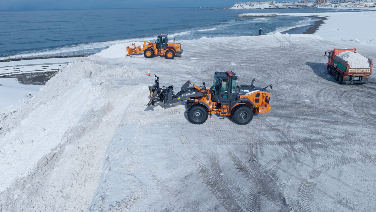 Two orange Hitachi snowplows and a truck clearing and loading snow near a snowy coastline.