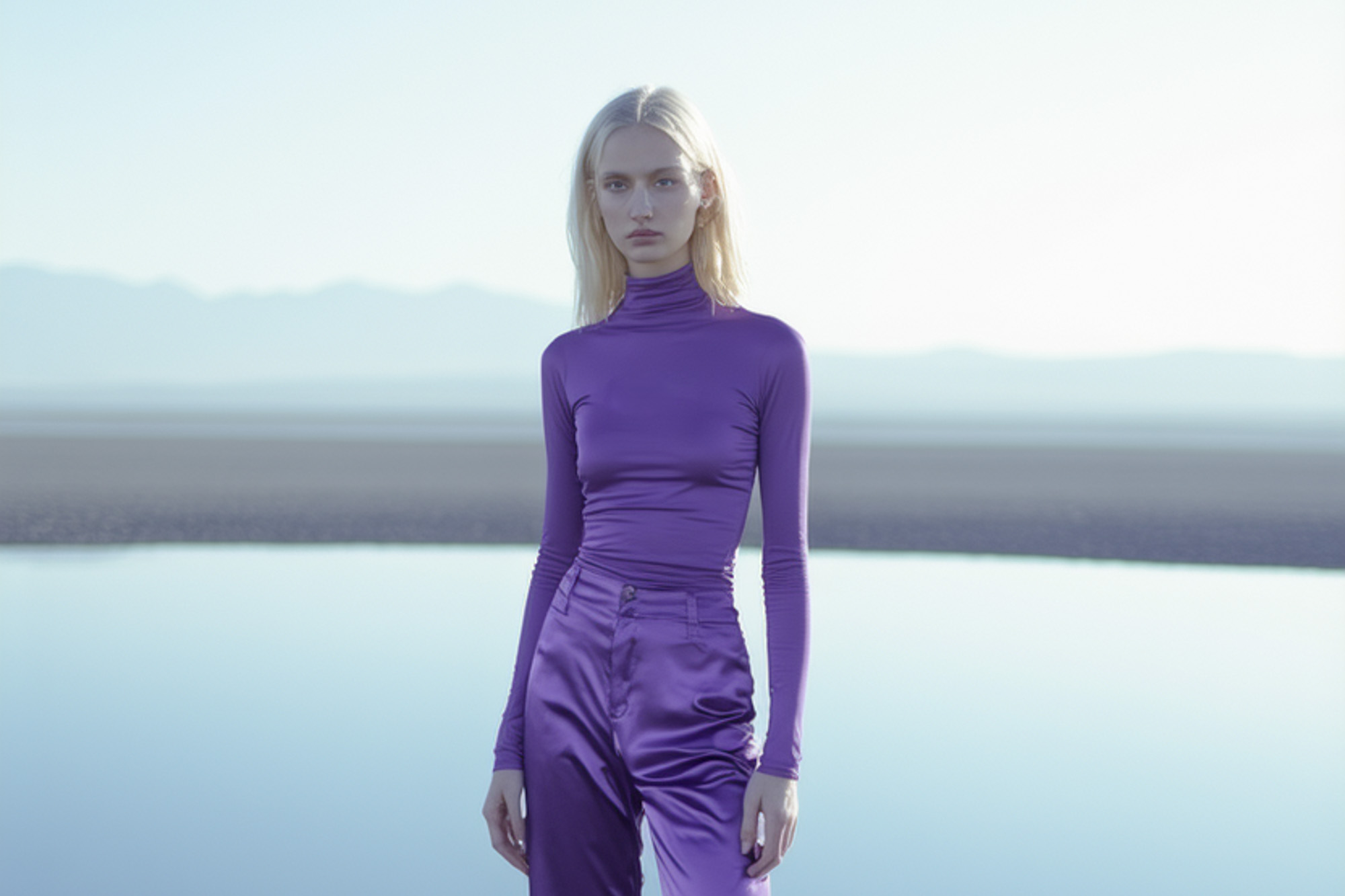 Blonde woman wearing a purple turtleneck and shiny purple pants standing in front of a calm body of water with mountains in the background.