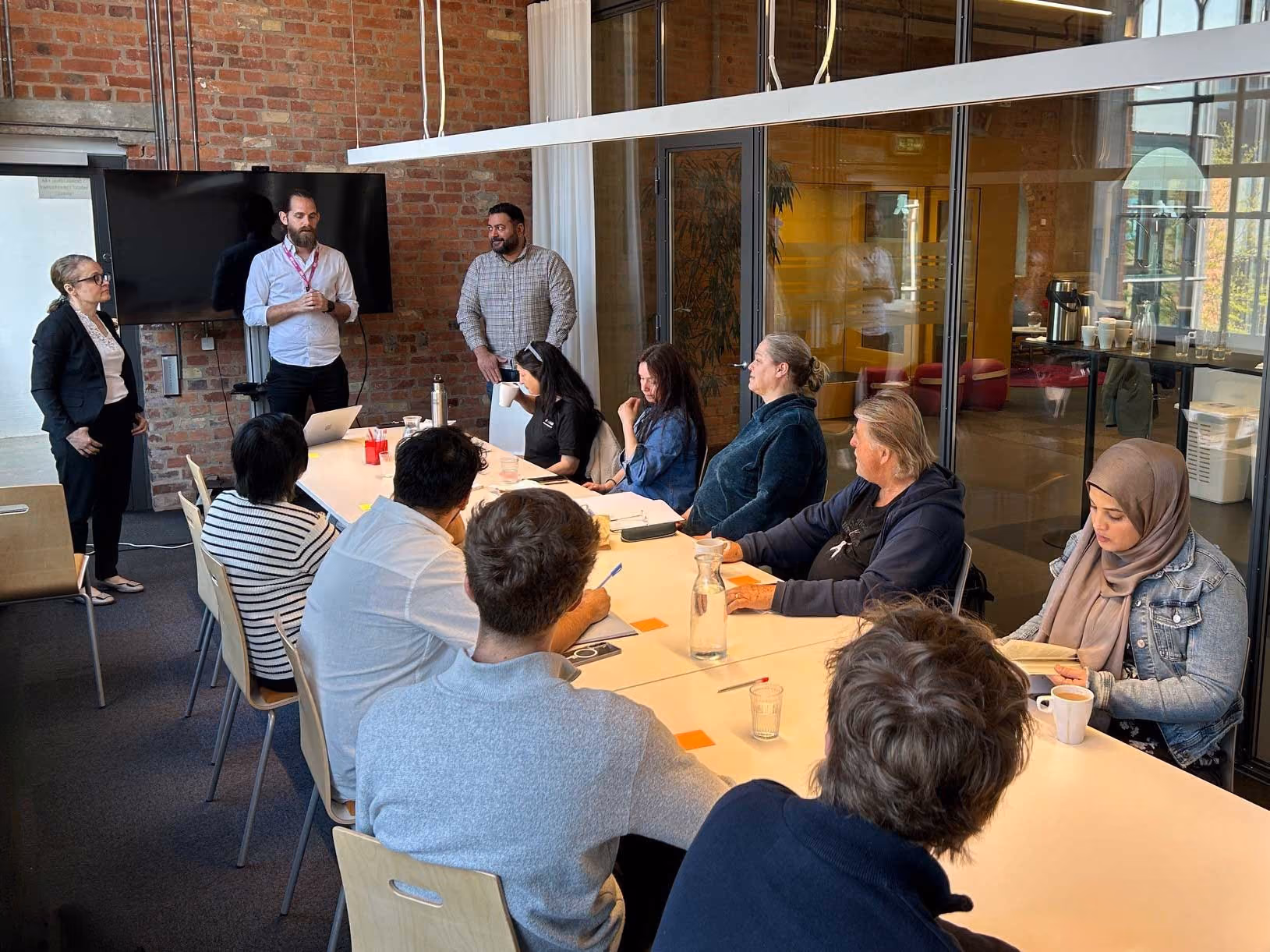 En grupp på tio personer deltar i en workshop i ett modernt konferensrum med tegelväggar.