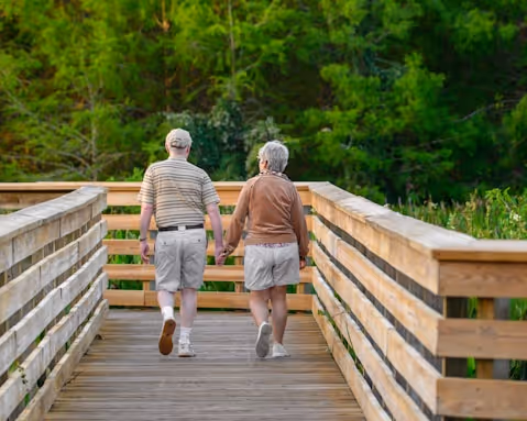 Older couple walking hand-in-hand on a wooden bridge surrounded by green nature.