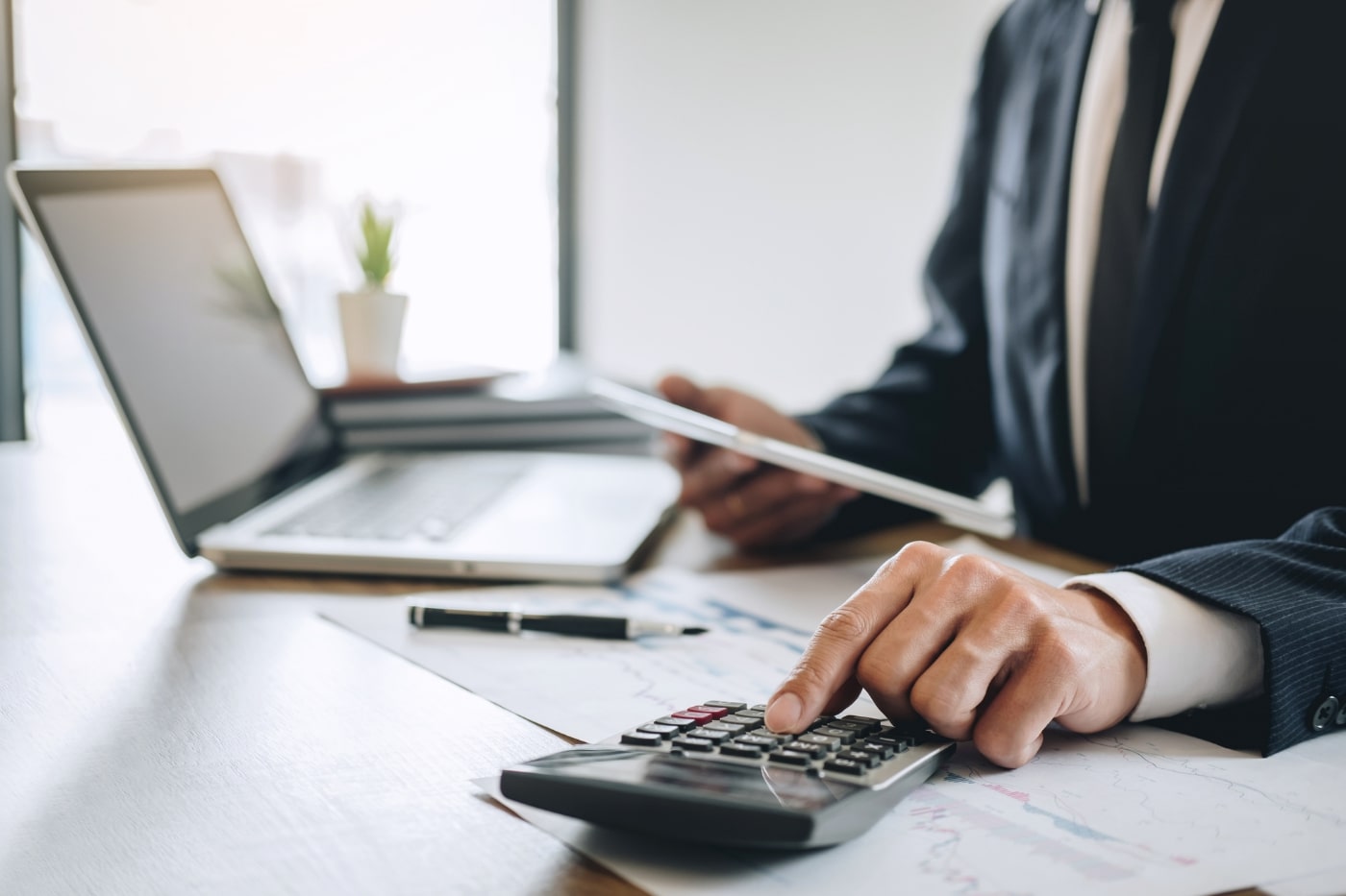 Business professional performing financial reconciliation using a calculator and reviewing financial documents on a desk with a laptop and tablet. Optimizing finance processes through precise reconciliation methods