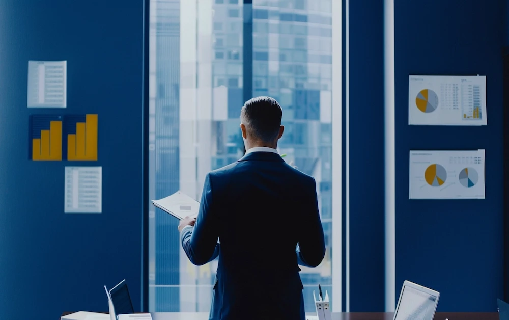 Financial analyst reviewing reports in an office with charts and graphs on the wall, representing the use of regulatory reporting software for accurate compliance and financial analysis.