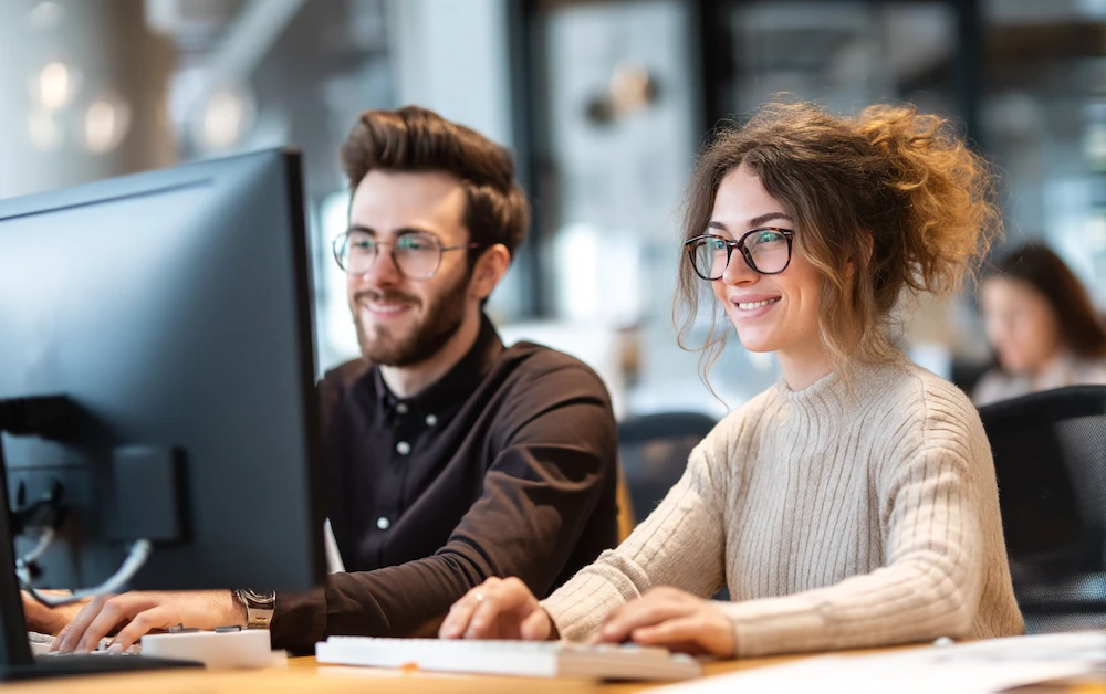 Two smiling accountants working together on adjusting entries at a computer, ensuring accurate period-end financial statements and compliance.
