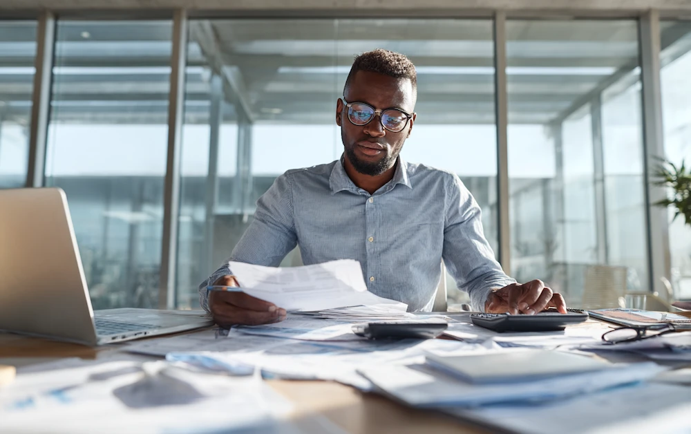 Finance professional analyzing prepaid expense reports at a desk with financial documents, calculator, and laptop in a modern office setting.