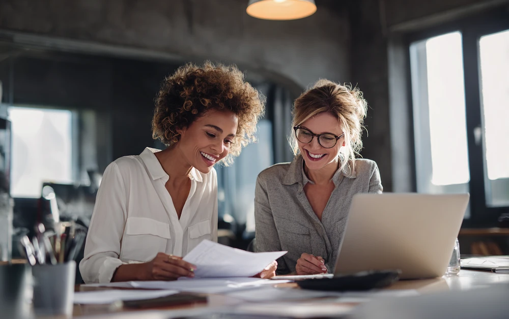 Two finance professionals smiling while reviewing documents and using a laptop to reconcile expenses in a modern office setting.