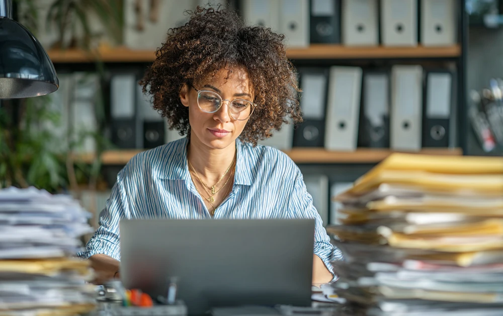 Finance professional managing the accounts receivable process on a laptop surrounded by stacks of invoices and documents, ensuring timely customer payments and accurate bookkeeping.