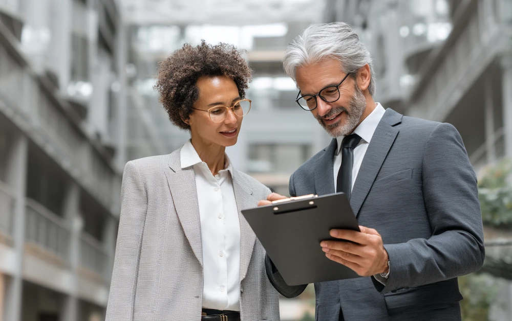 Two business professionals reviewing governance risk and compliance (GRC) documentation on a clipboard in a modern corporate office setting