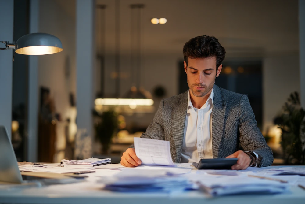 A focused finance professional in a modern office works late at night, reviewing paper invoices and using a calculator, surrounded by piles of documents — illustrating the manual processes that accounts receivable automation can streamline for greater efficiency and accuracy.