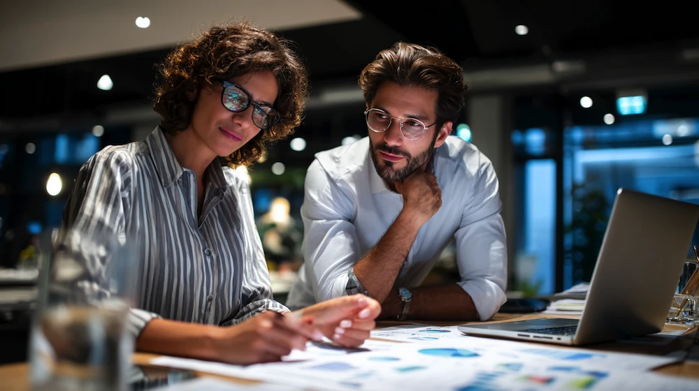 Two finance professionals reviewing compliance reporting data and financial charts in a modern office, analyzing accuracy and regulatory performance for corporate reporting.