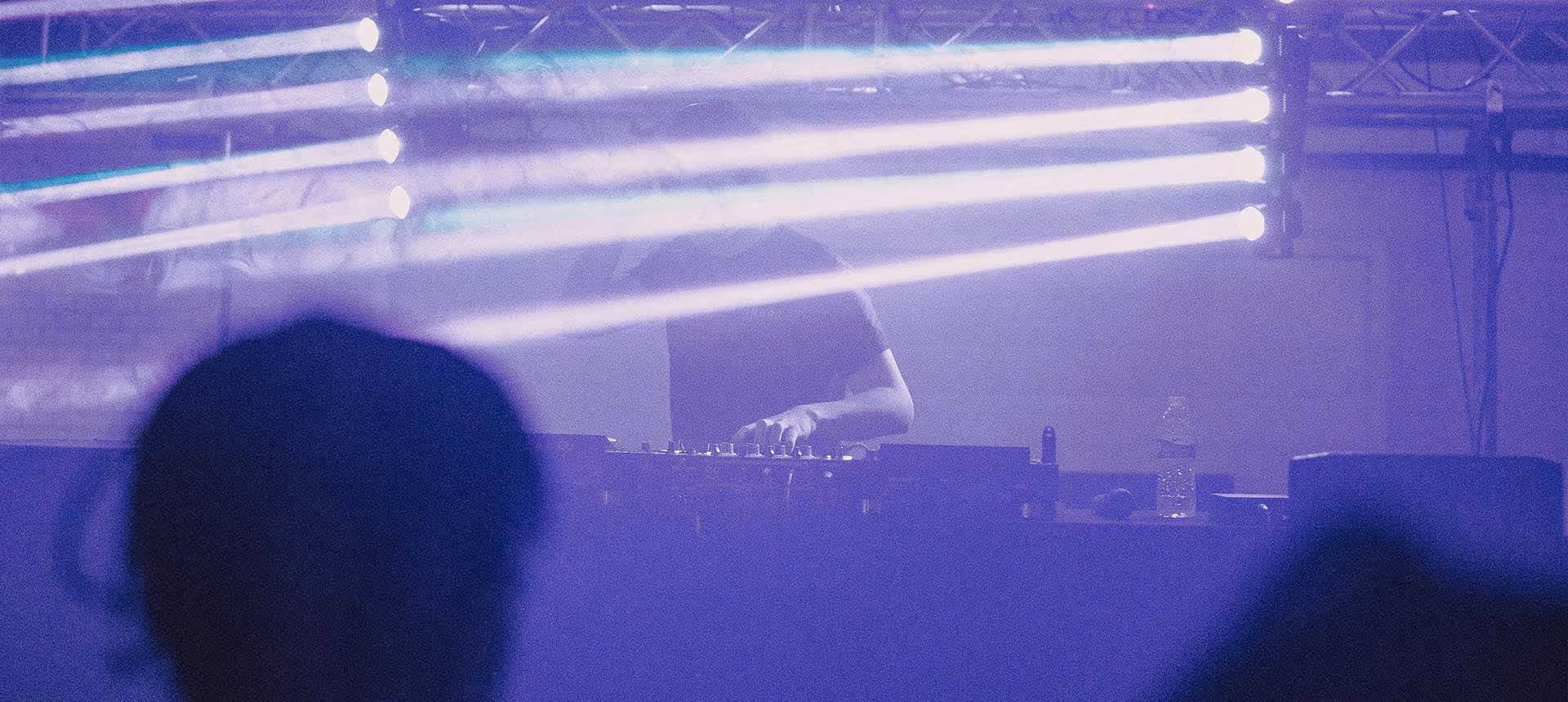 Ein DJ legt in einem vernebeltem Club auf, schemenhaft tanzen menschen auf der Tanzfläche