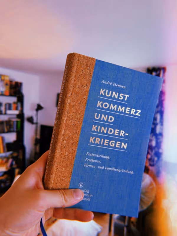 André Hennens Buch "Kunst Kommerz und Kinderkriegen" wird von einer Hand hochgehalten.