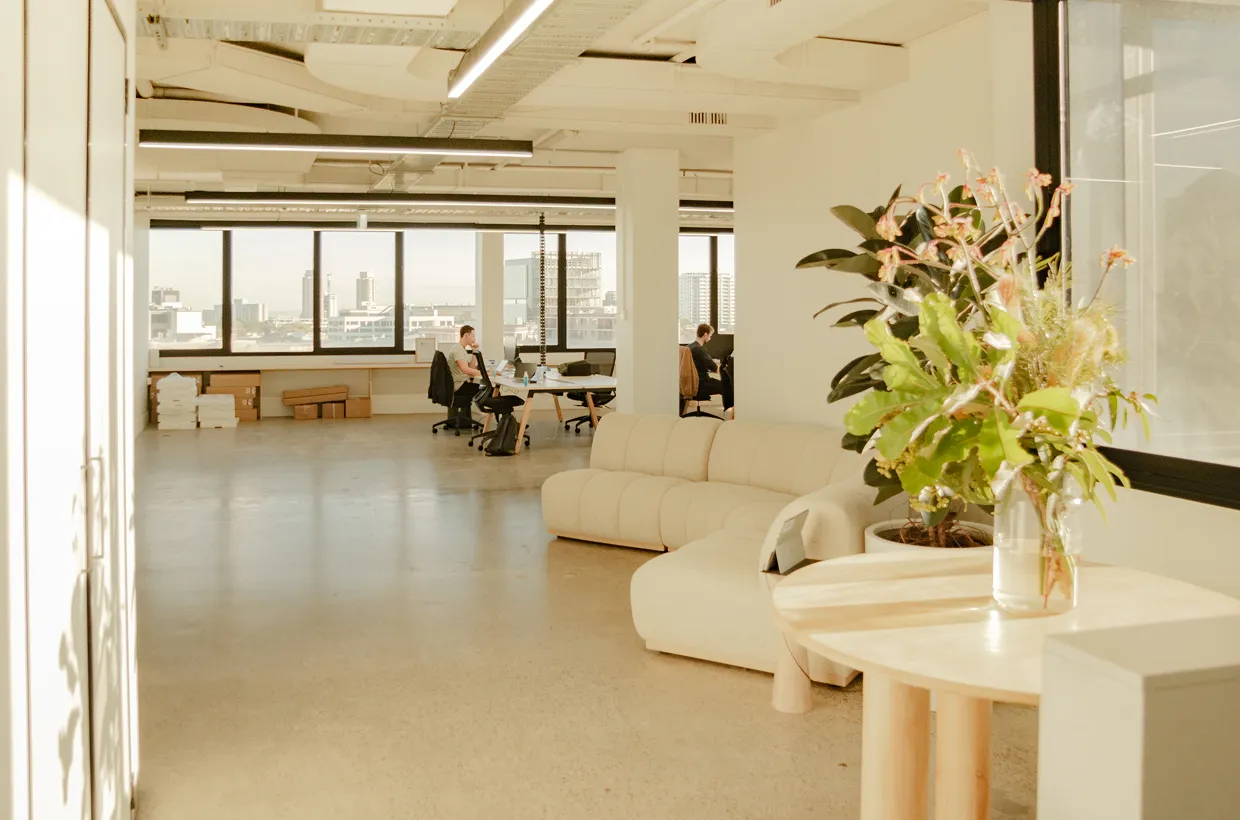 Modern open-plan office with large windows offering city views, minimalistic furniture including a white sectional sofa and a round wooden table with a glass vase of fresh flowers in the foreground. A few people are working at desks in the background.