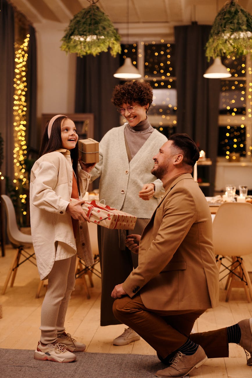 A family celebrating Christmas in a cozy, decorated living room with warm lighting and festive decor. A father kneels to give a gift to his daughter, while the mother smiles with another gift in hand. Christmas lights twinkle in the background, creating a joyful holiday atmosphere.