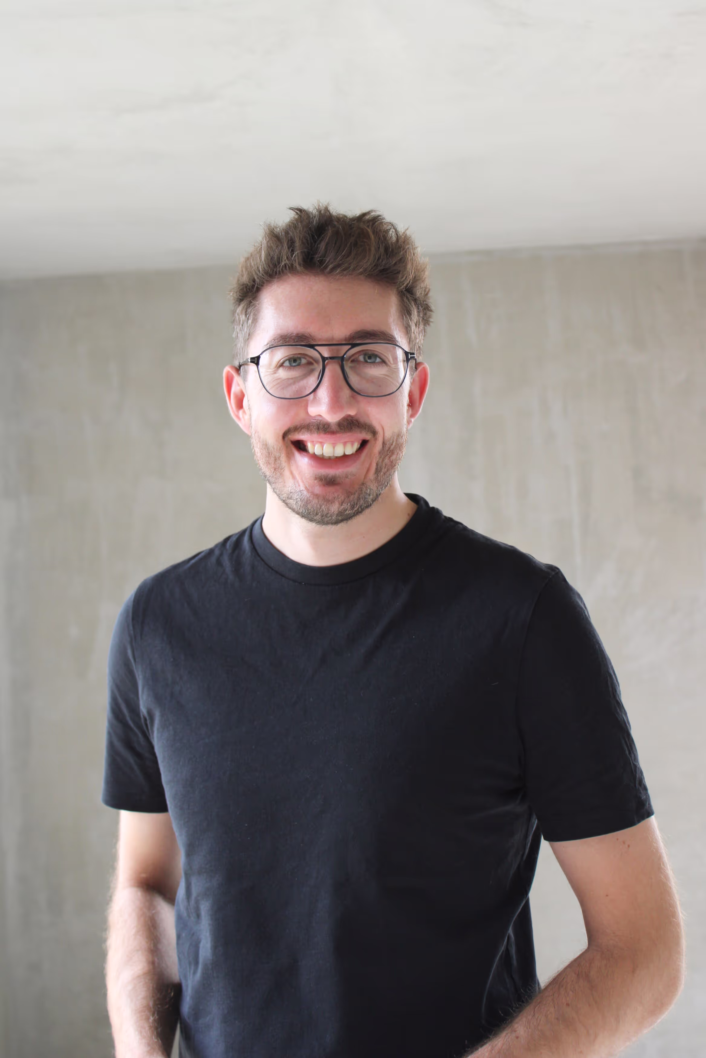 Smiling man with glasses and beard wearing a black t-shirt standing against a gray wall.