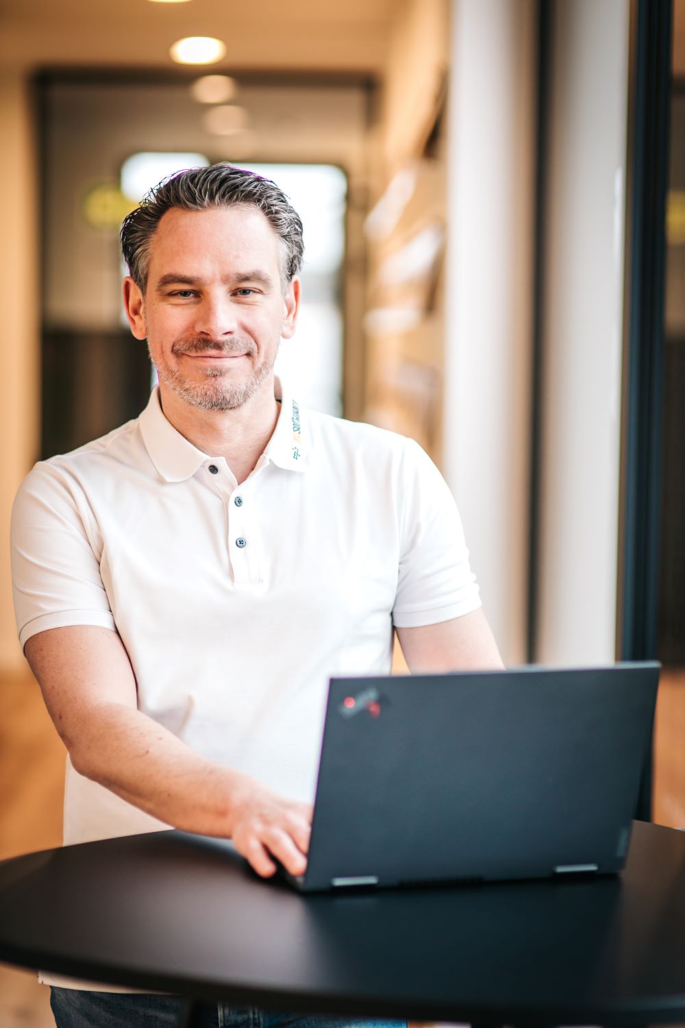 Man in a white polo shirt smiling while using a laptop at a black round table indoors.