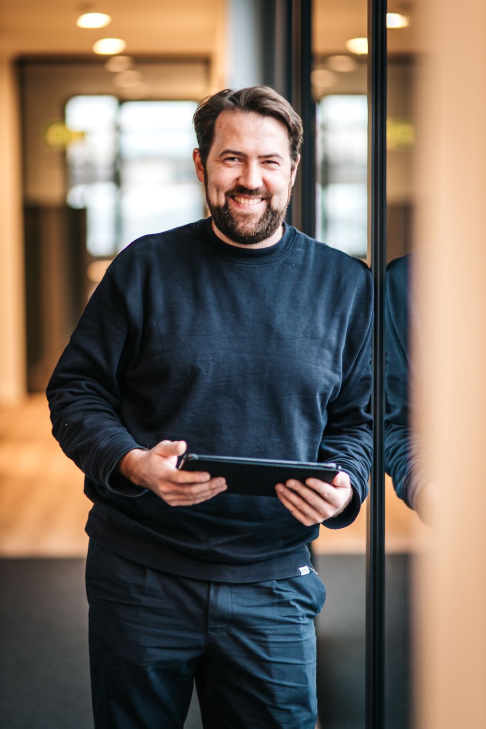 Smiling man with beard leaning against glass wall holding a tablet in a modern office.