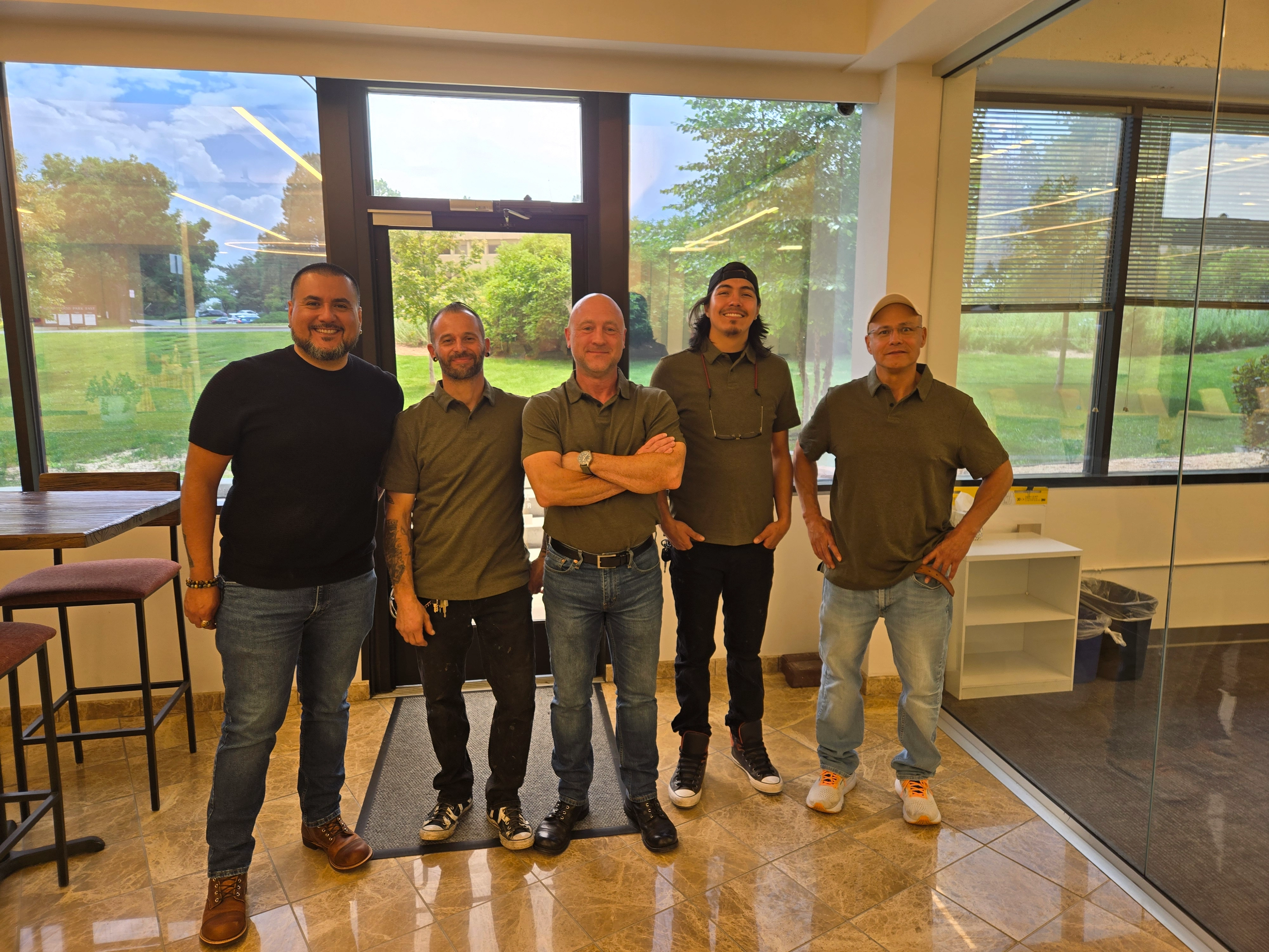 Five men stand inside an office with a glass wall, smiling. Sunlight streams in, highlighting their casual attire.