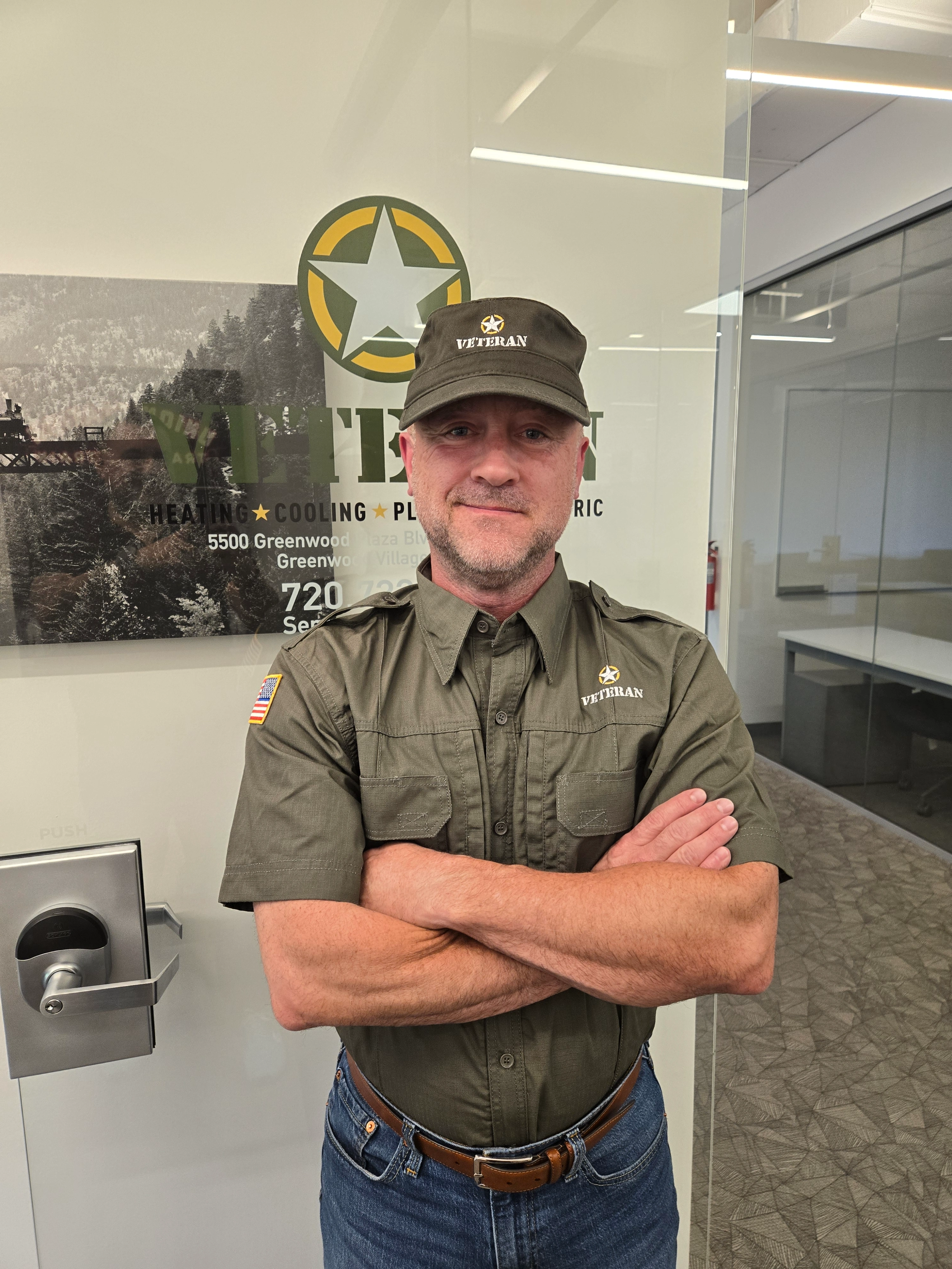 Man in a green "Veteran" uniform and cap stands confidently with arms crossed