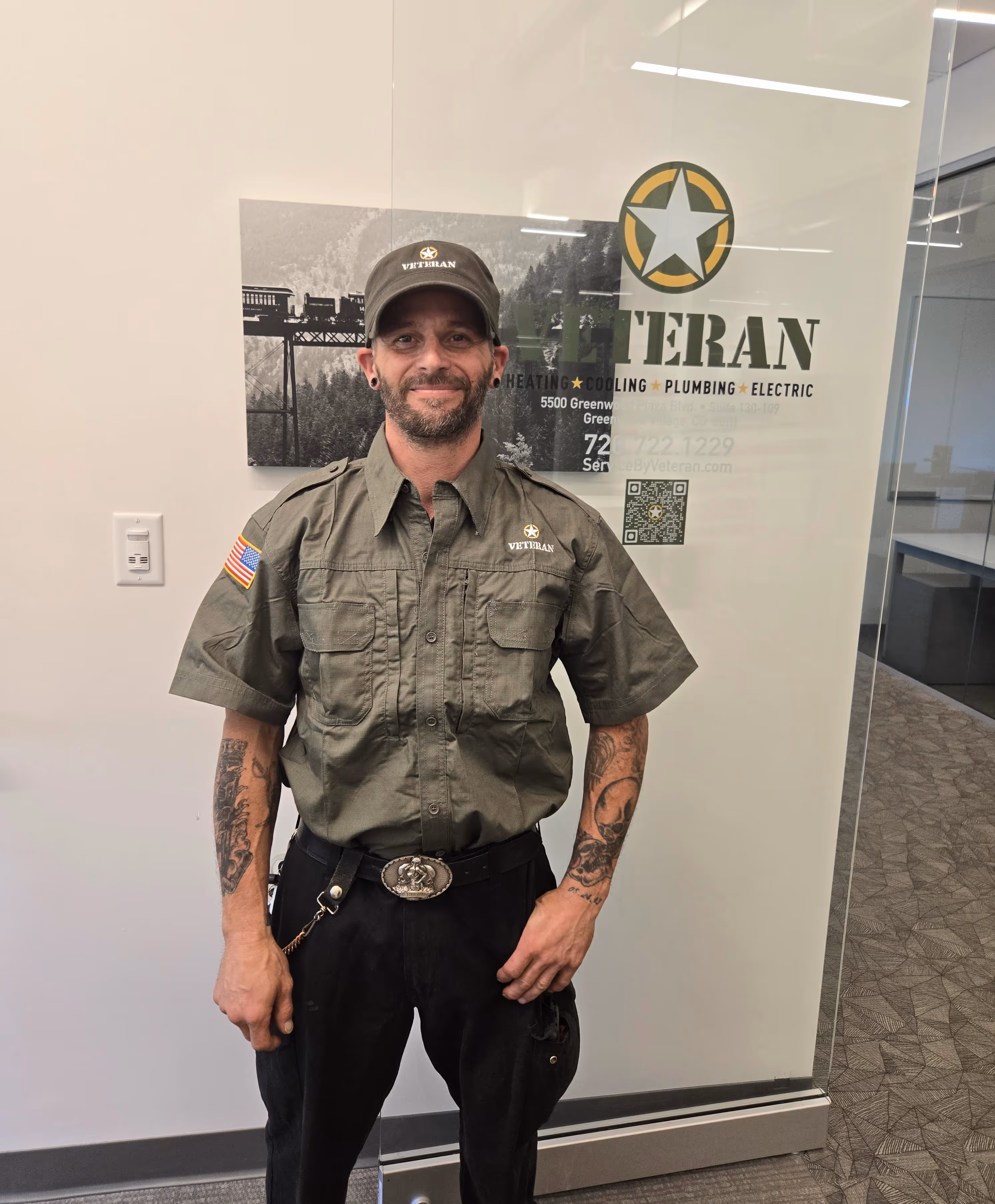 A man in a "Veteran" branded olive uniform and cap stands smiling in an office. 