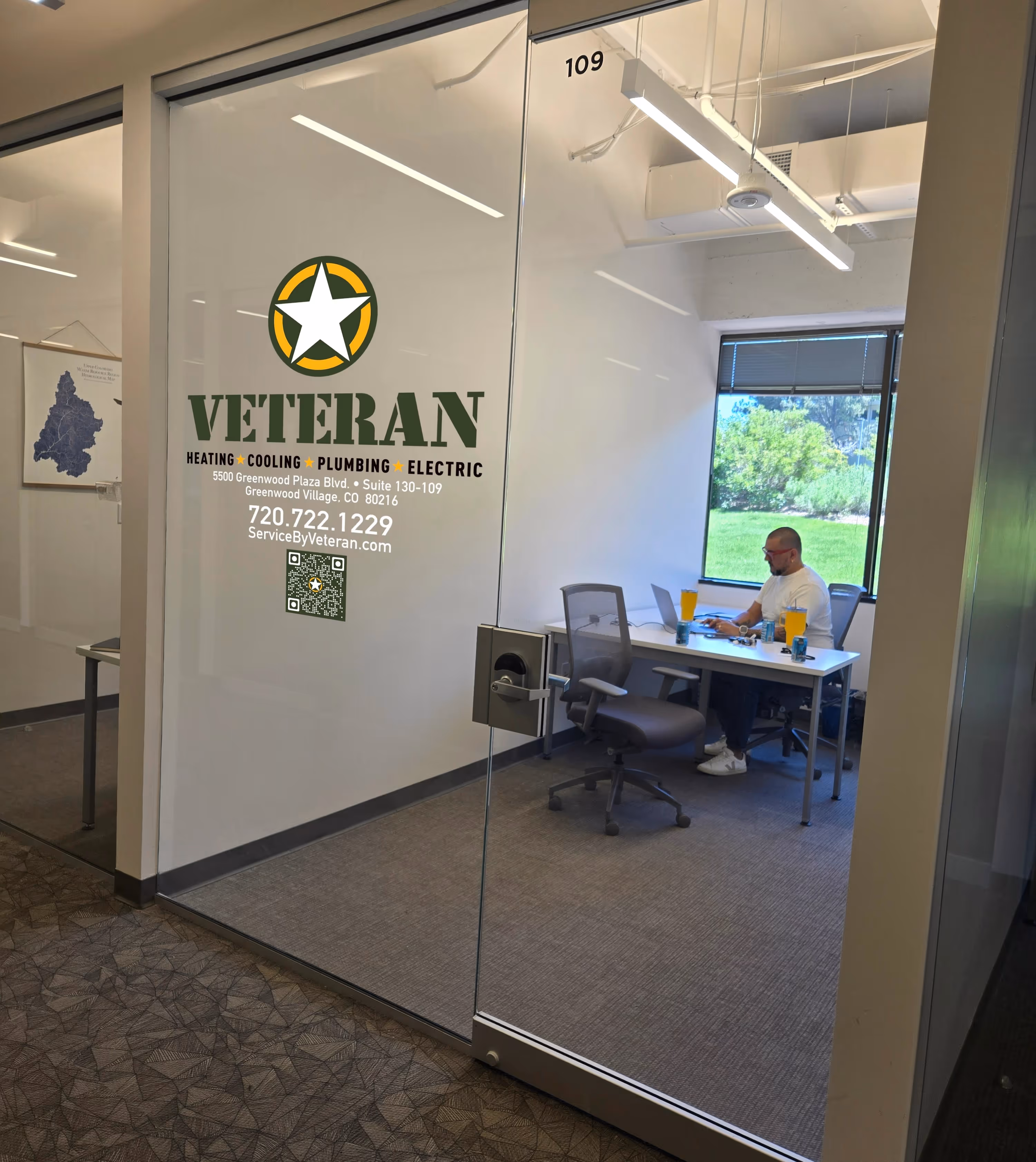 A man sits at a white desk in a bright, modern office labeled "Veteran Heating Cooling Plumbing Electric." 