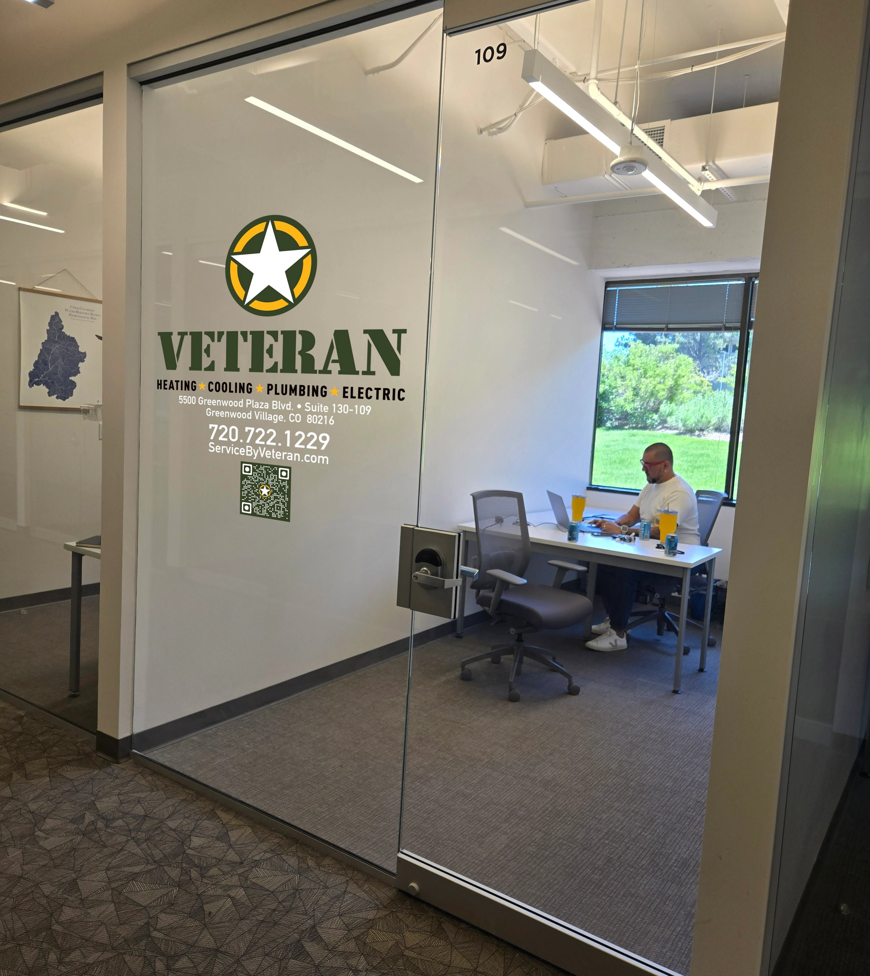 Glass office door with "Veteran Heating Cooling Plumbing Electric" logo. Inside, a person works at a desk with a laptop, window view of greenery.