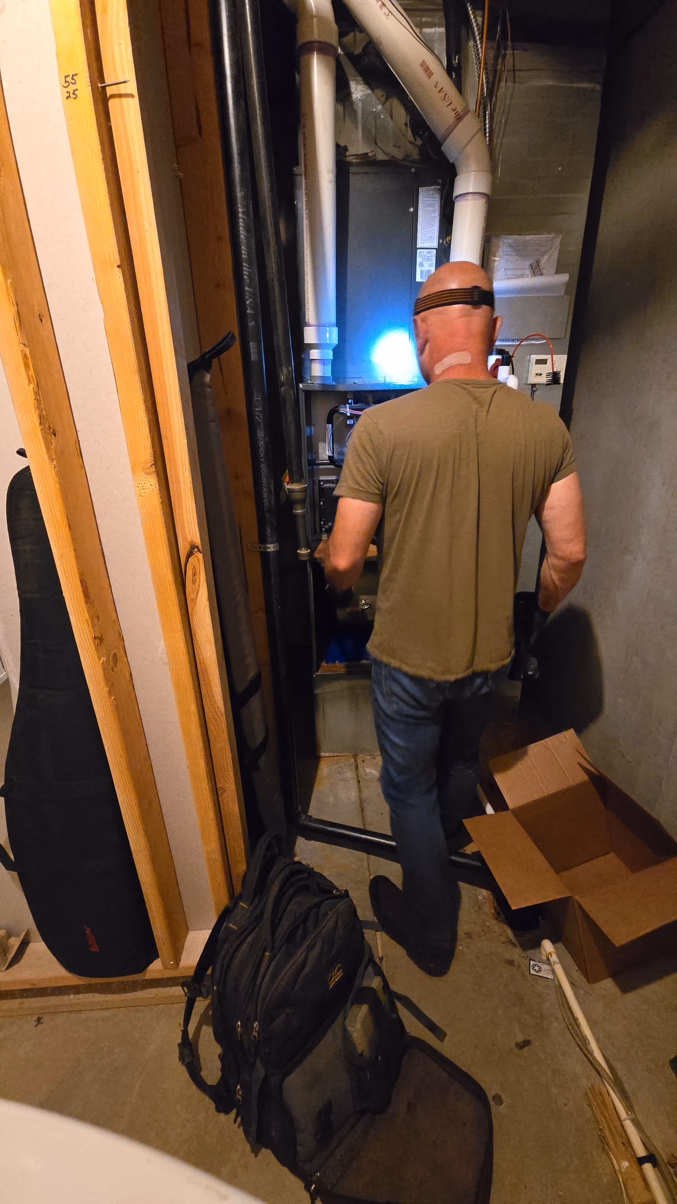 A man wearing a headlamp works on a furnace in a dimly lit utility room. 