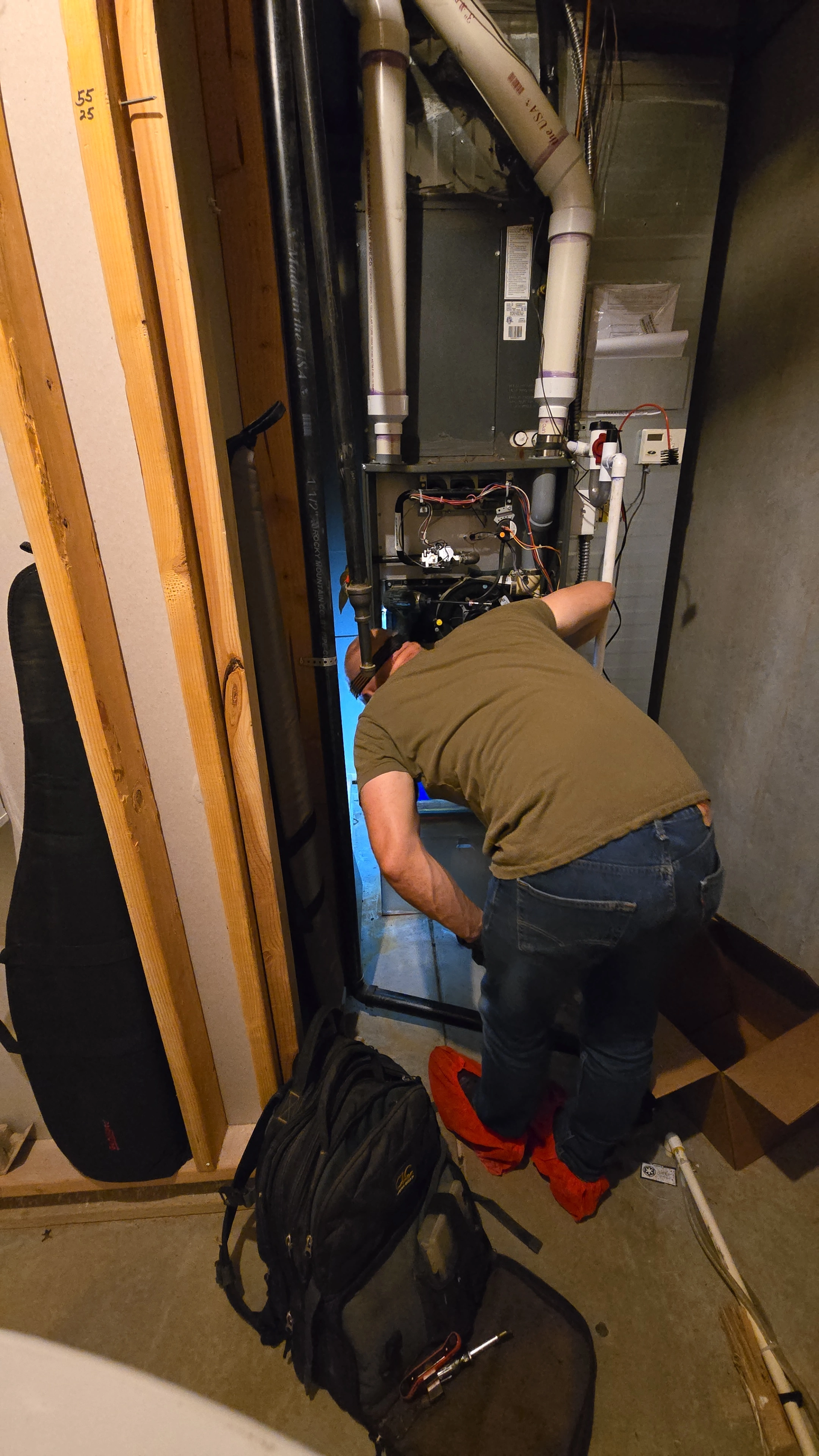 A person in a green shirt and red shoe covers inspects a furnace in a dimly lit basement.