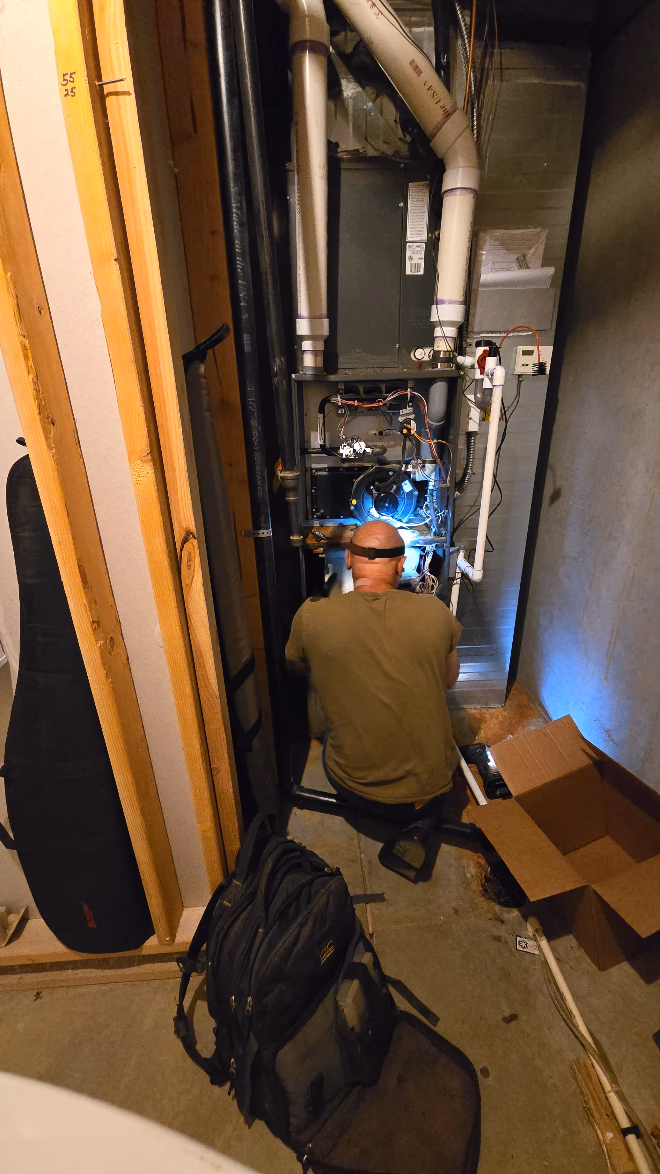 A man wearing glasses crouches in front of an exposed furnace in a dimly lit basement, surrounded by wires and pipes, focused on repairs.