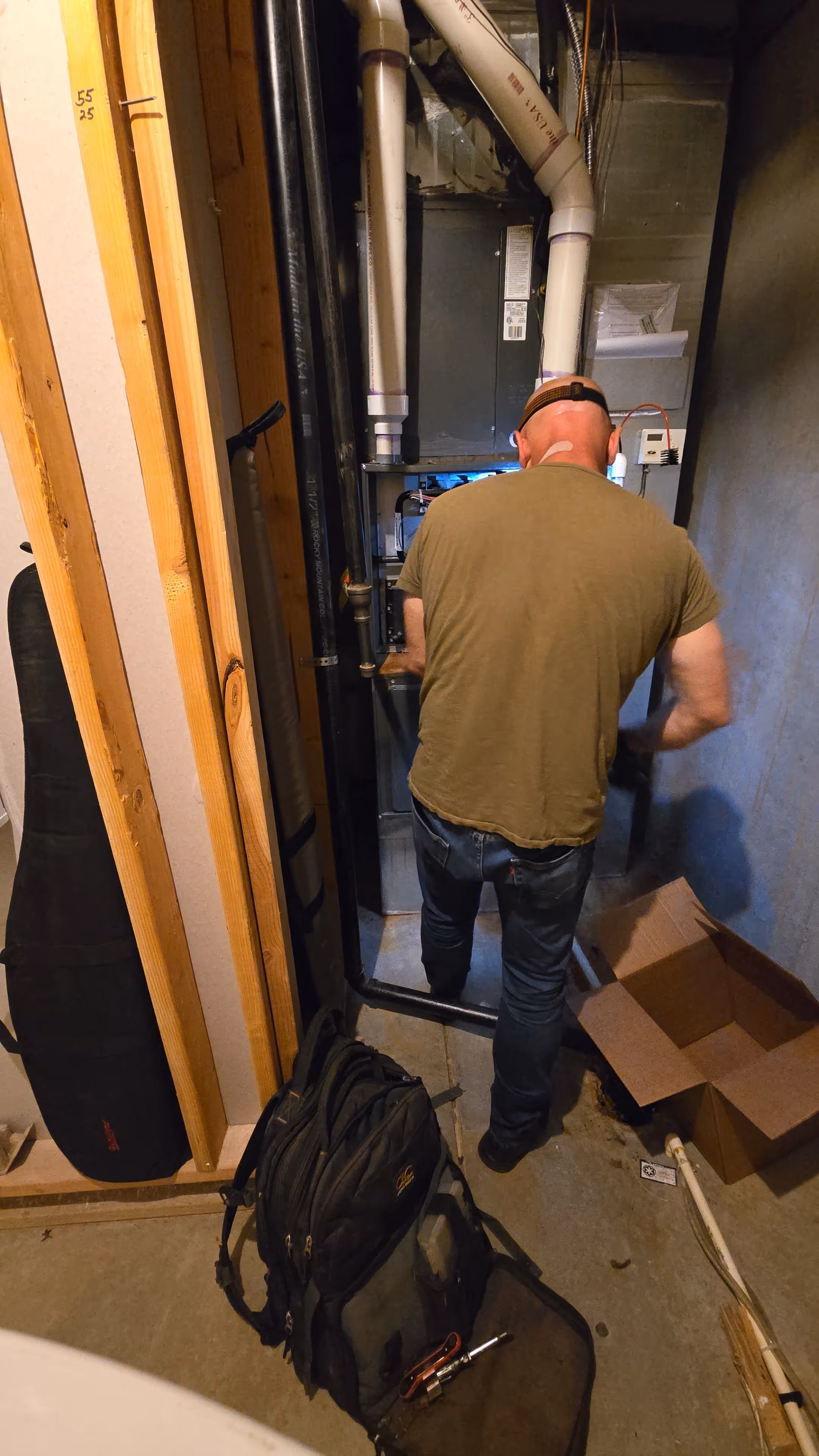 A man wearing glasses crouches in front of an exposed furnace in a dimly lit basement, surrounded by wires and pipes, focused on repairs.