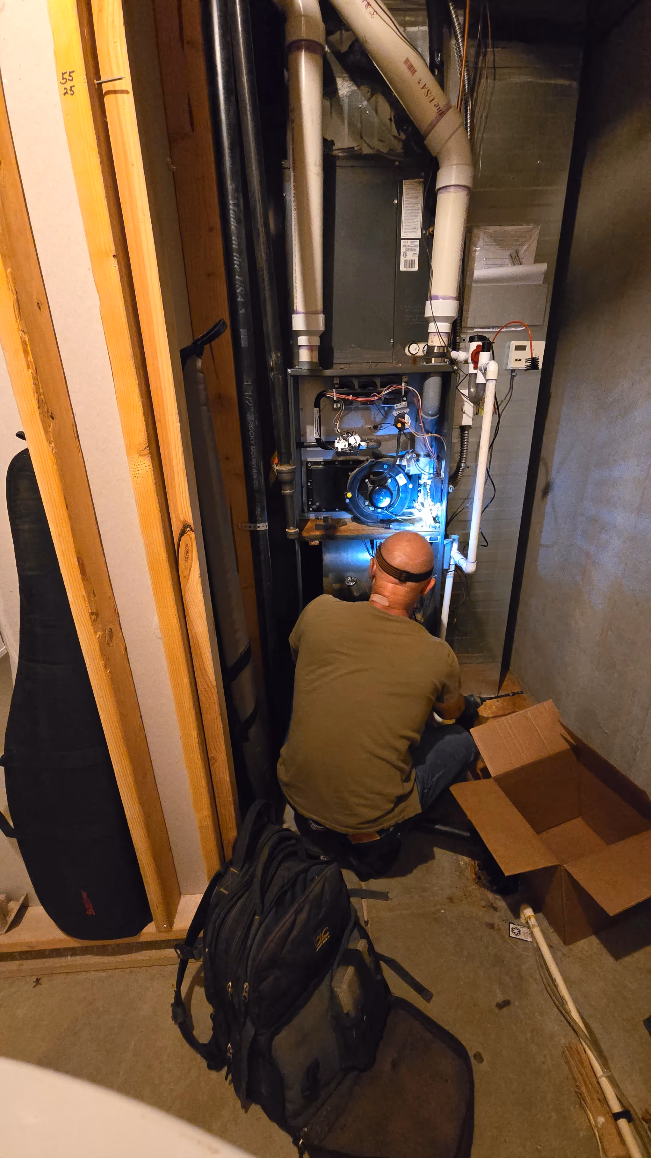 A man wearing glasses crouches in front of an exposed furnace in a dimly lit basement, surrounded by wires and pipes, focused on repairs.