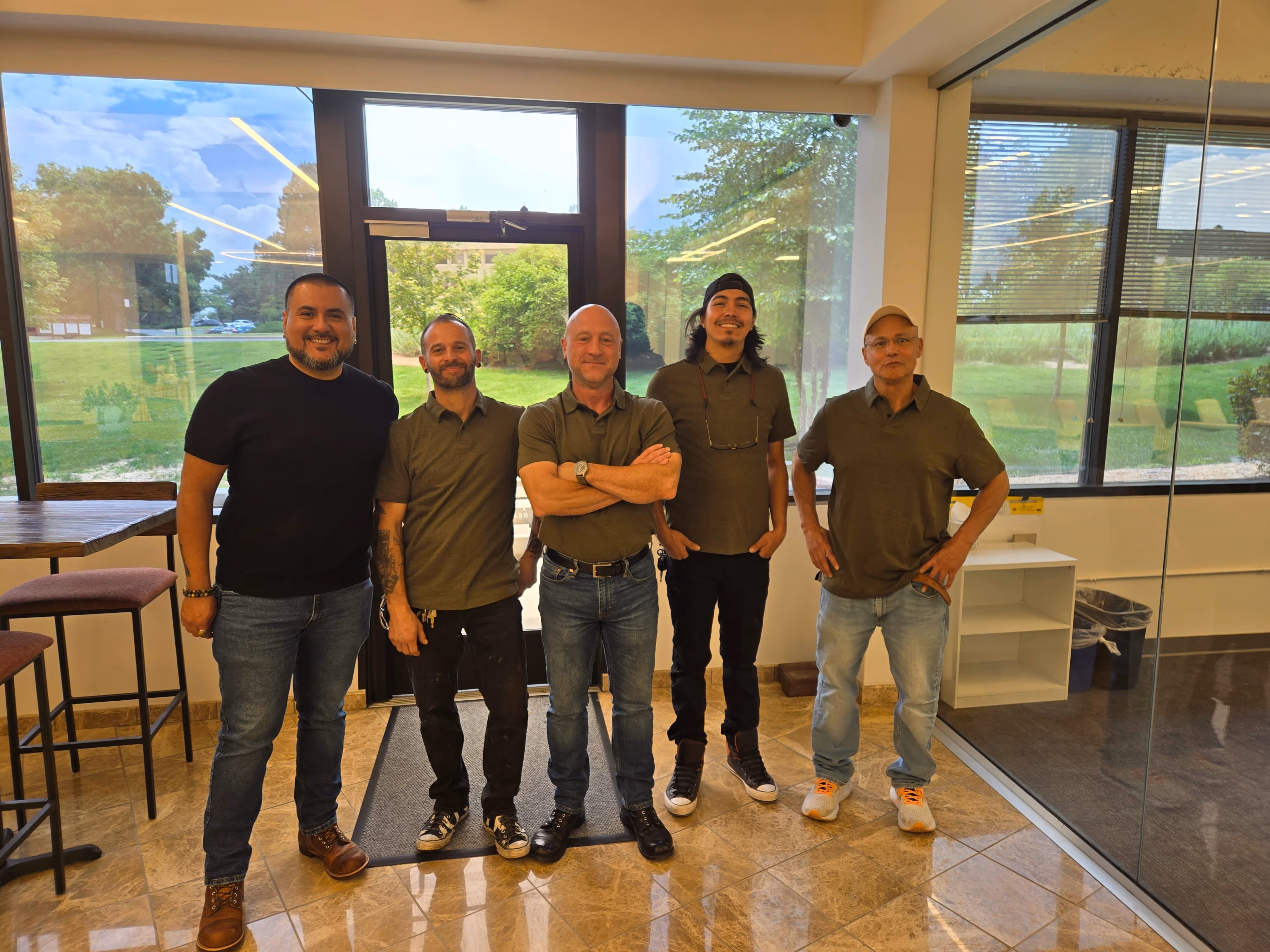Five men standing in a bright room with large windows showing a green outdoor view.