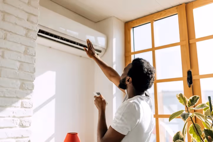 A person reaching up towards a wall-mounted mini-split air conditioner, holding a remote control.