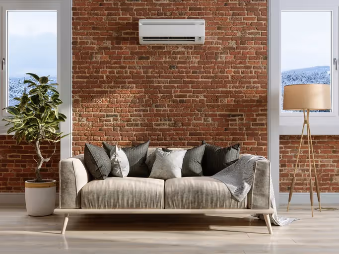 A living room with a mini-split air conditioner mounted above a sofa against a brick wall, flanked by two windows and a potted plant and a lamp.