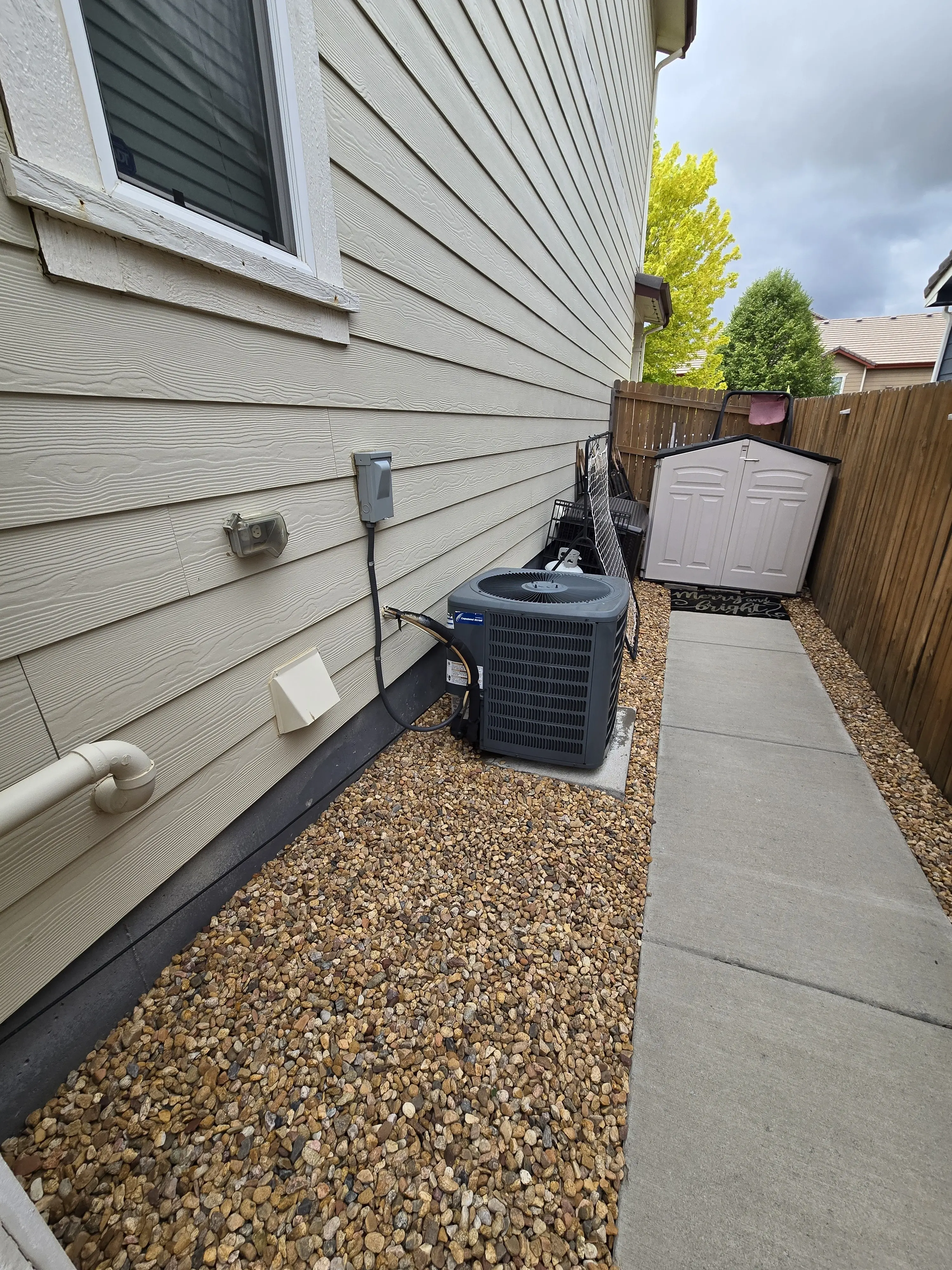 Side of a house with wooden siding and a small window. An air conditioning unit sits on gravel next to the wall.