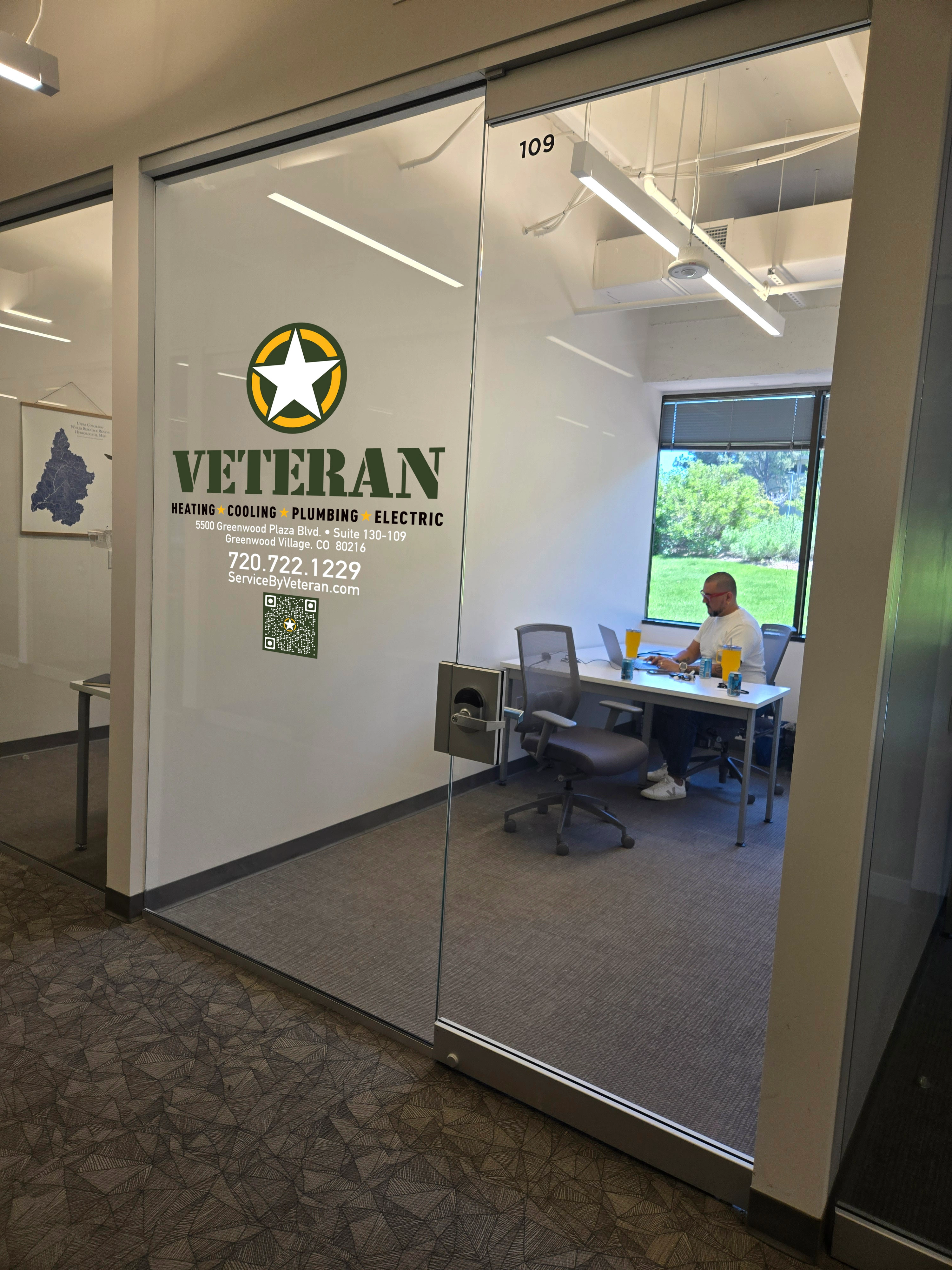 Office with glass door shows a logo for "Veteran" services, featuring heating, cooling, plumbing, and electric. Inside, a man works at a desk