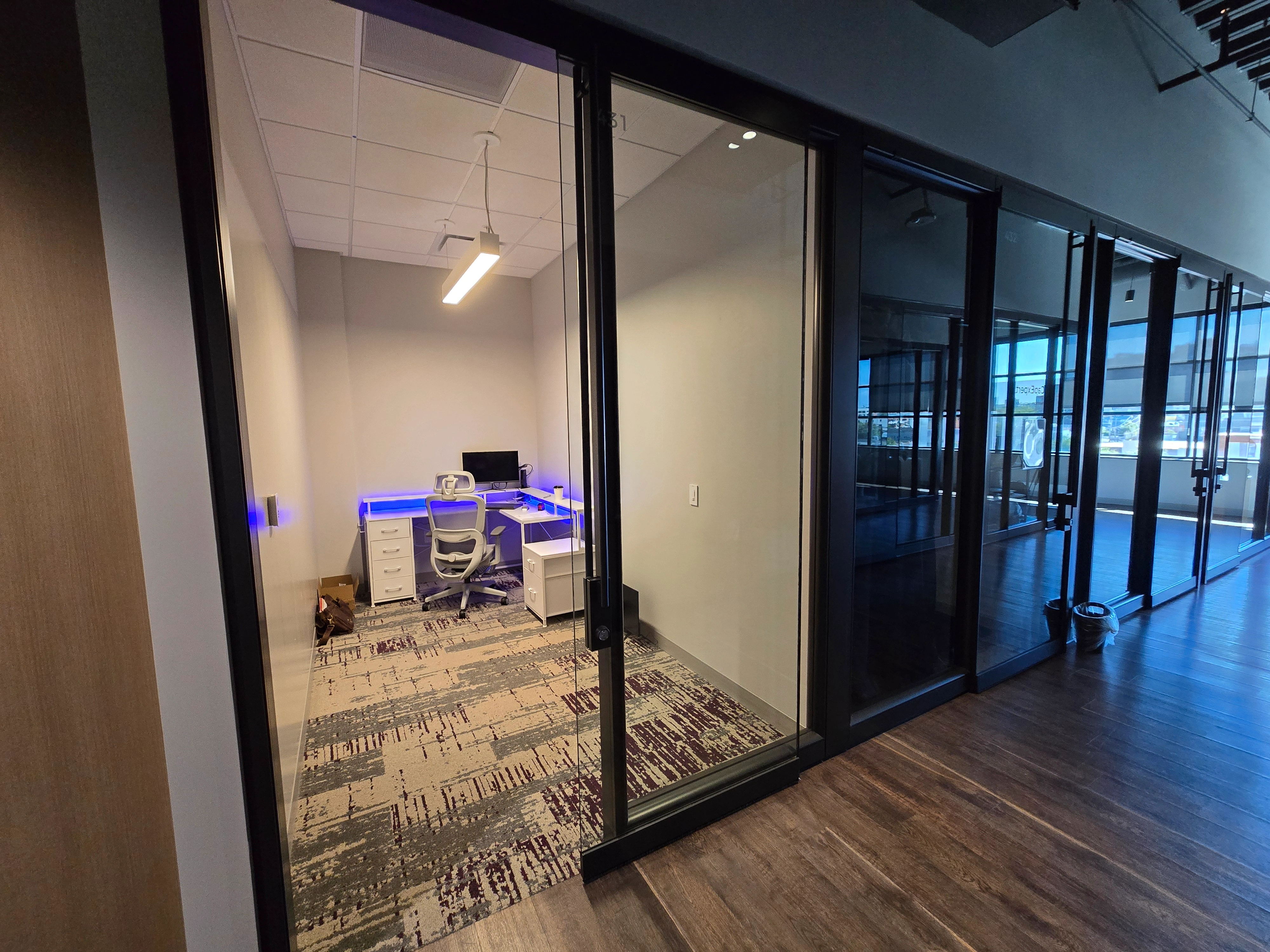 Un cubículo de oficina moderno con paredes de vidrio, con un escritorio con una computadora y iluminación de acento azul.