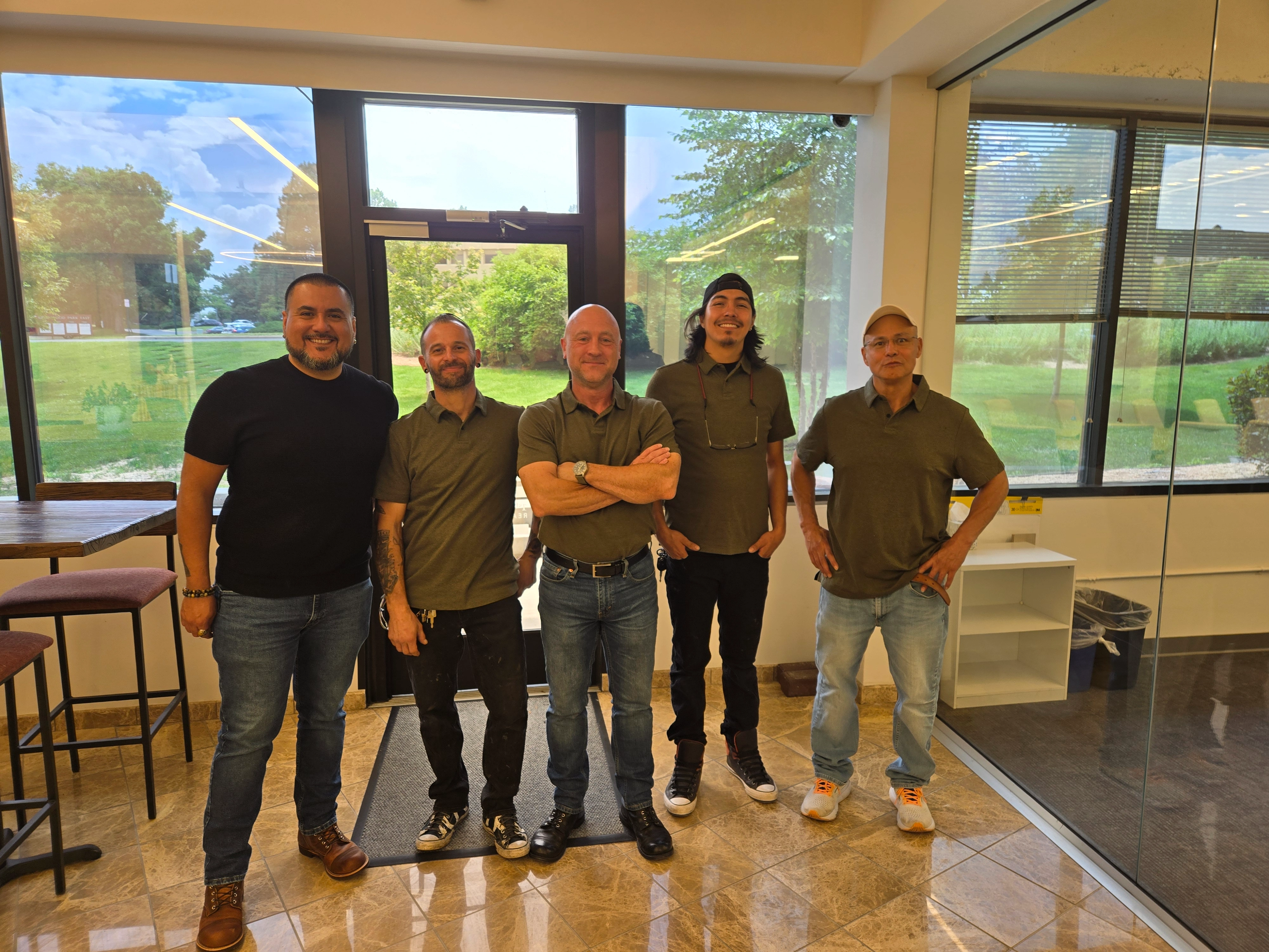 Cinco hombres de pie en una habitación brillante con grandes ventanas que muestran una vista verde al aire libre.